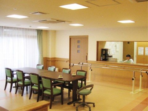四角い長机が２台並べられ、その周りには緑色の椅子が置いてある。入口ドア近くには、歩行訓練用の手すりが設置されている。