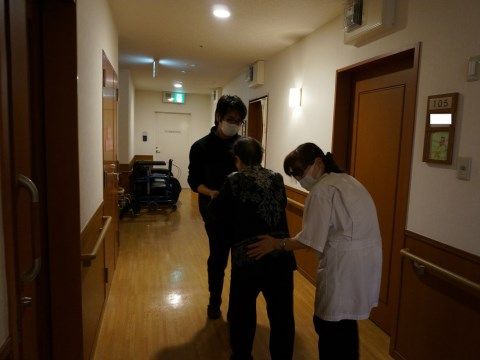 廊下で、男性スタッフに手を取られ、女性スタッフに後ろを支えらながら、歩行訓練を行う女性入居者
