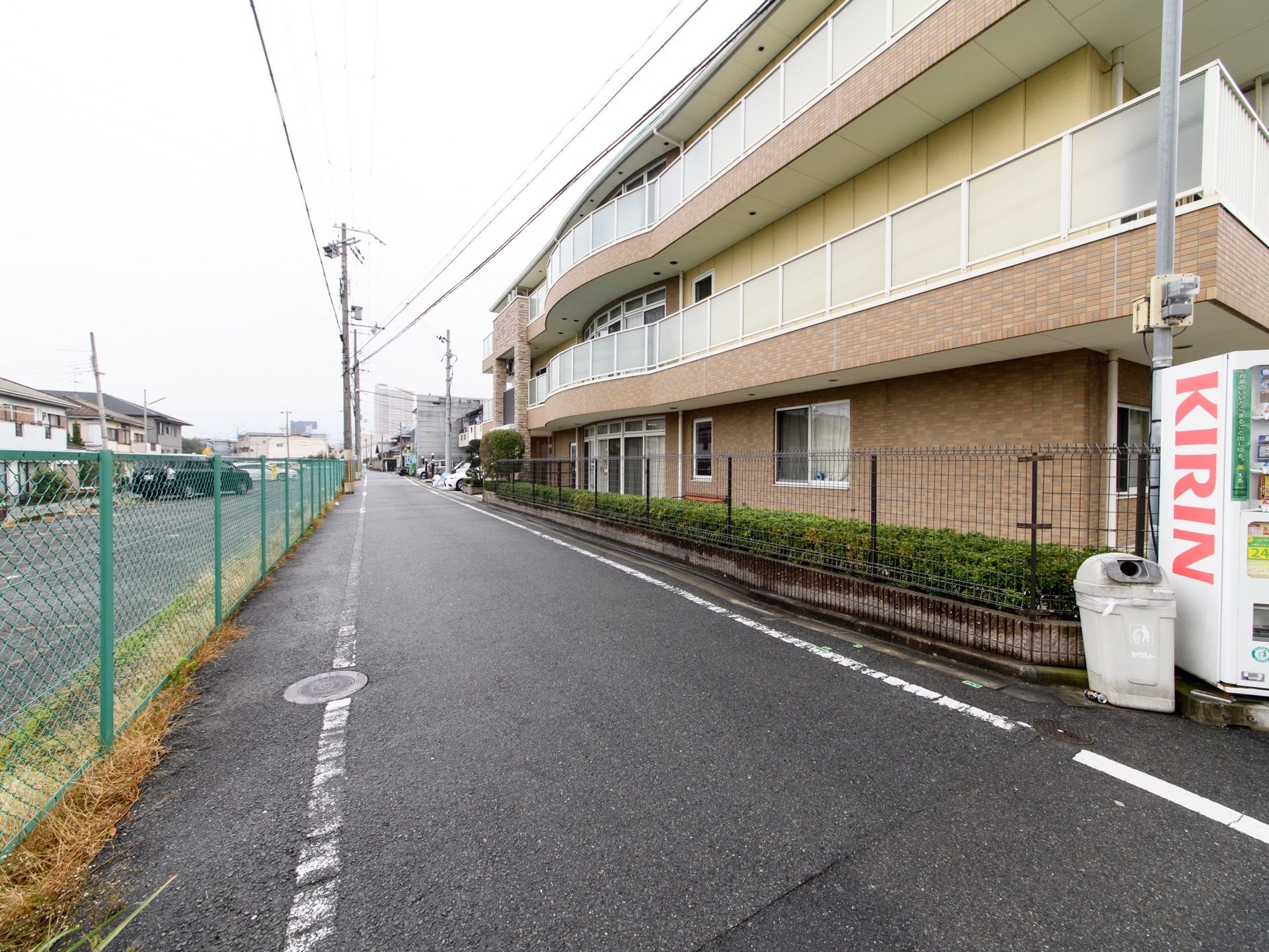 きれいに舗装されている道路である。三階建てで落ち着いた色の建物である。施設前には自動販売機が設置されている。