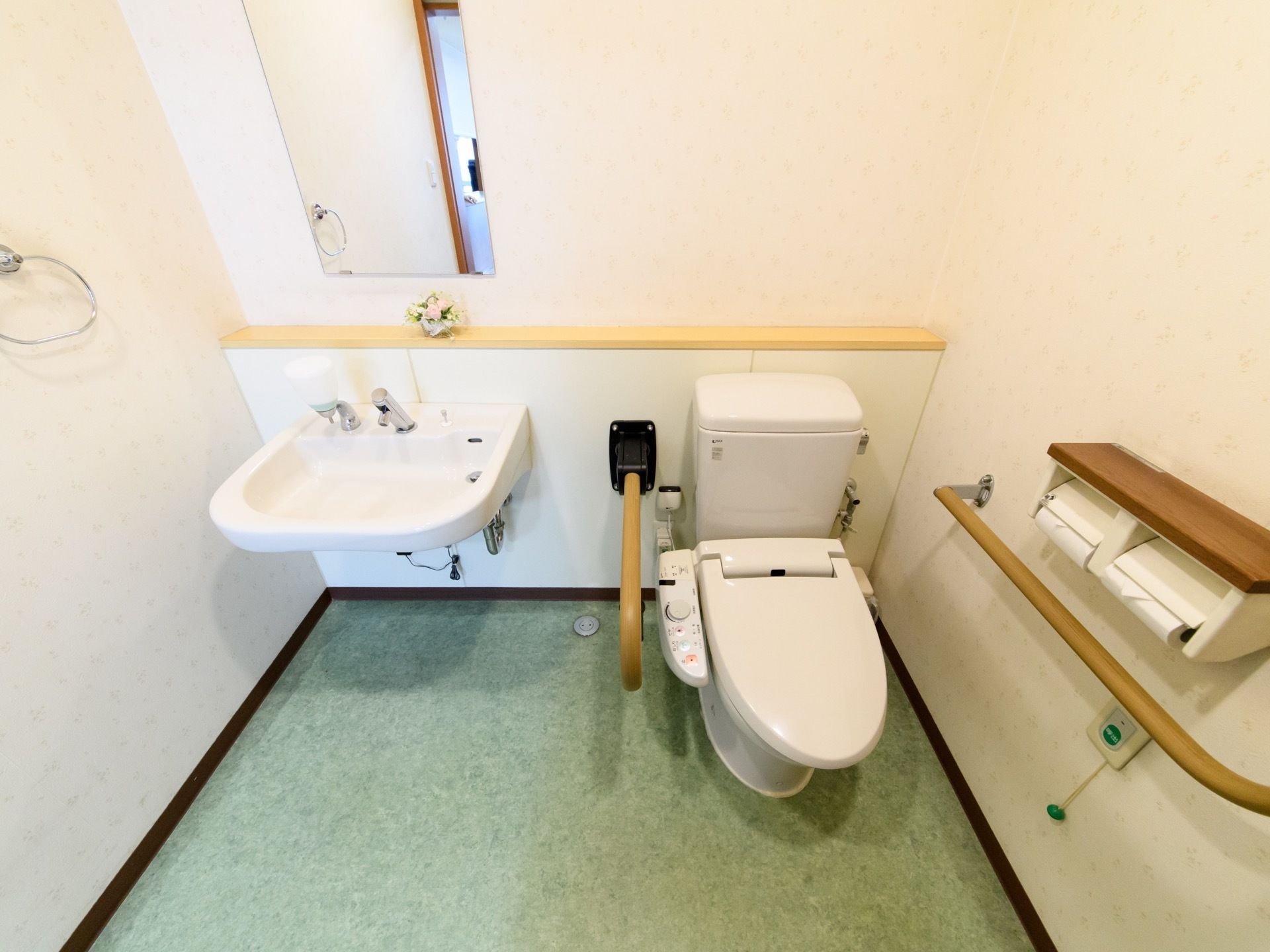 トイレには手すりがついた温水洗浄便座と並んで足元は空間になっている洗面ボウルがある。便座の横にペーパーホルダーと非常ボタンがある。