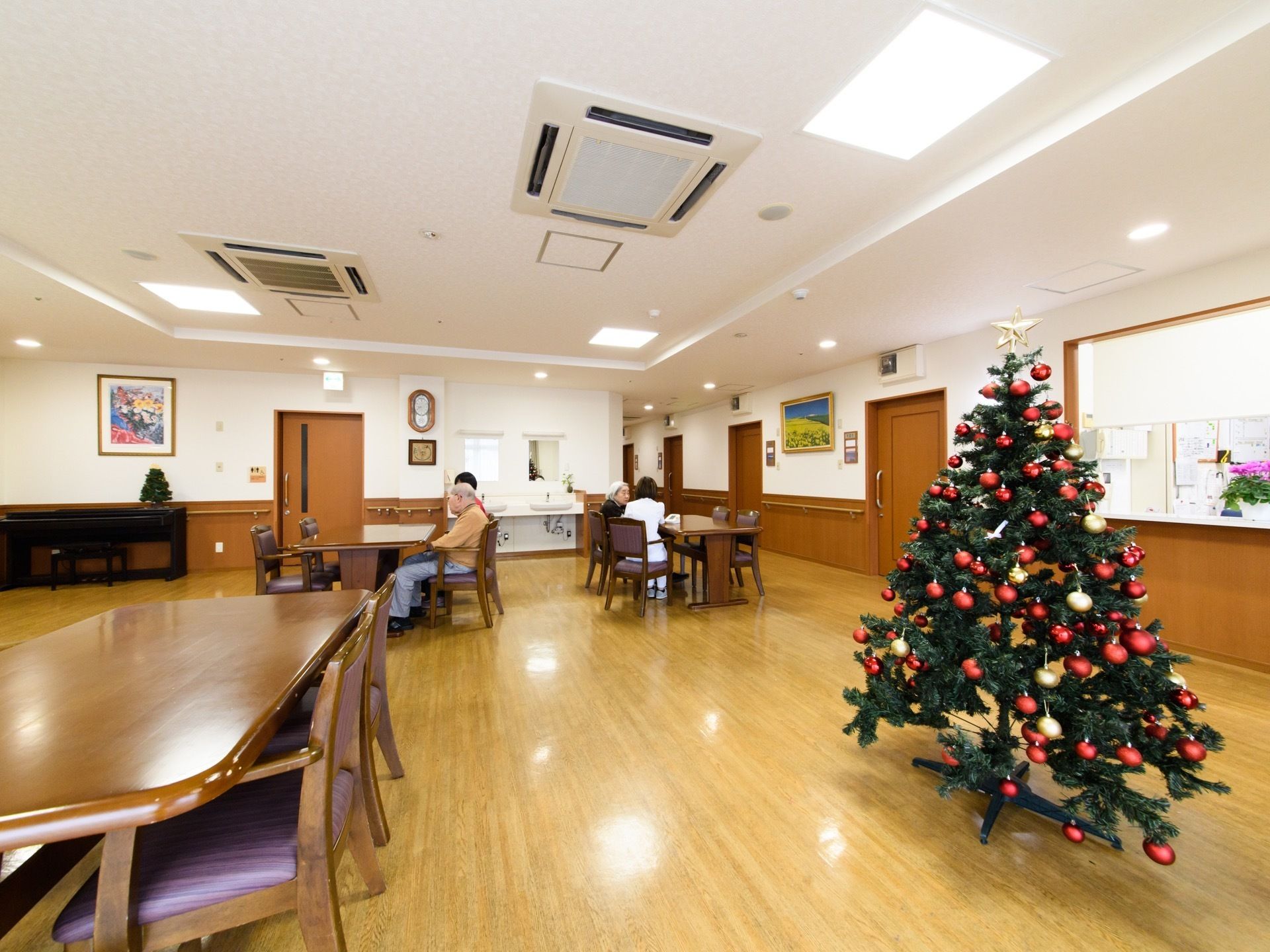 各居室前の通路のつづきに共有スペースがある。クリスマスツリーが飾られている。４人掛けテーブルが間隔をあけて置かれている。