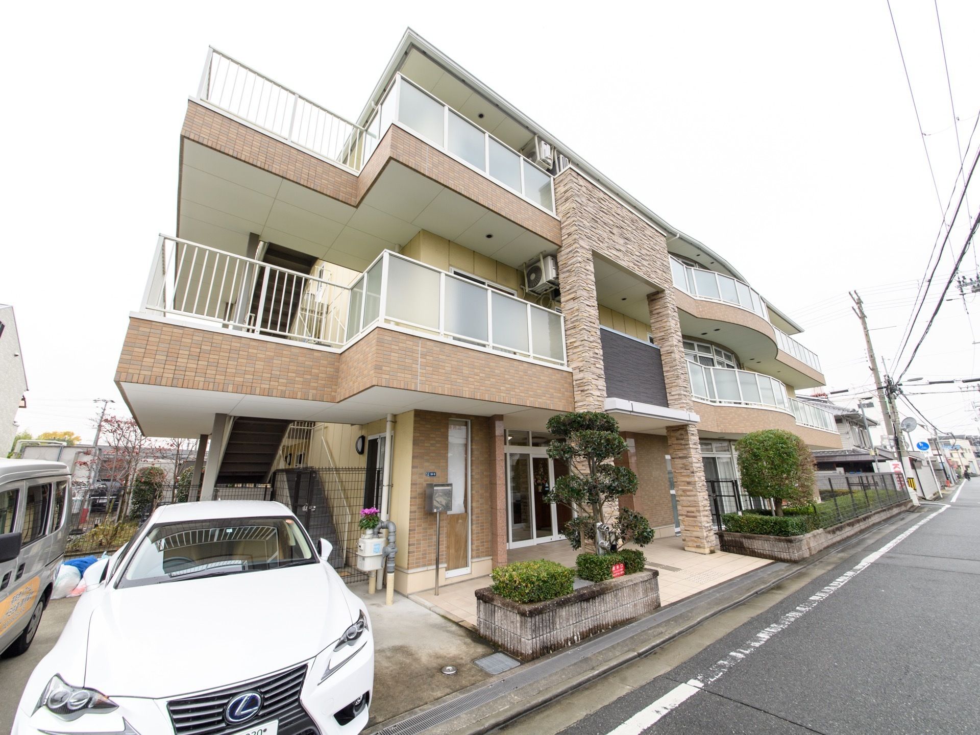 道路に沿って３階建ての施設が建っている。各階に周回するようにベランダある。１階道路沿いに植栽がある。
