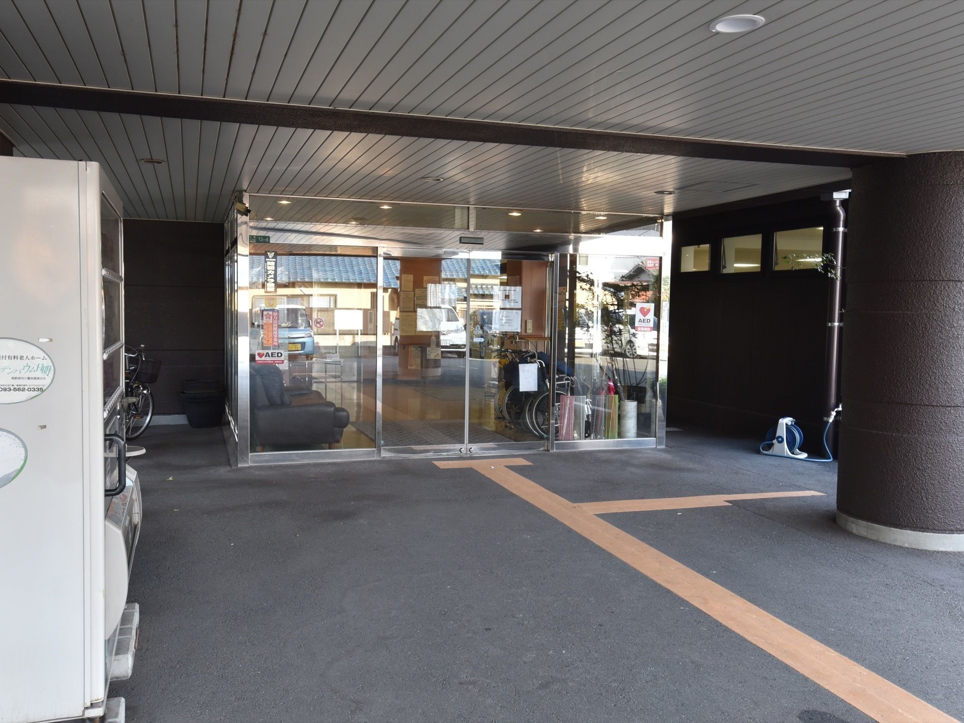 玄関入口前のスペースである。横側には自動販売機を設置している。ドアの端の部分にAEDのシールを貼っている。