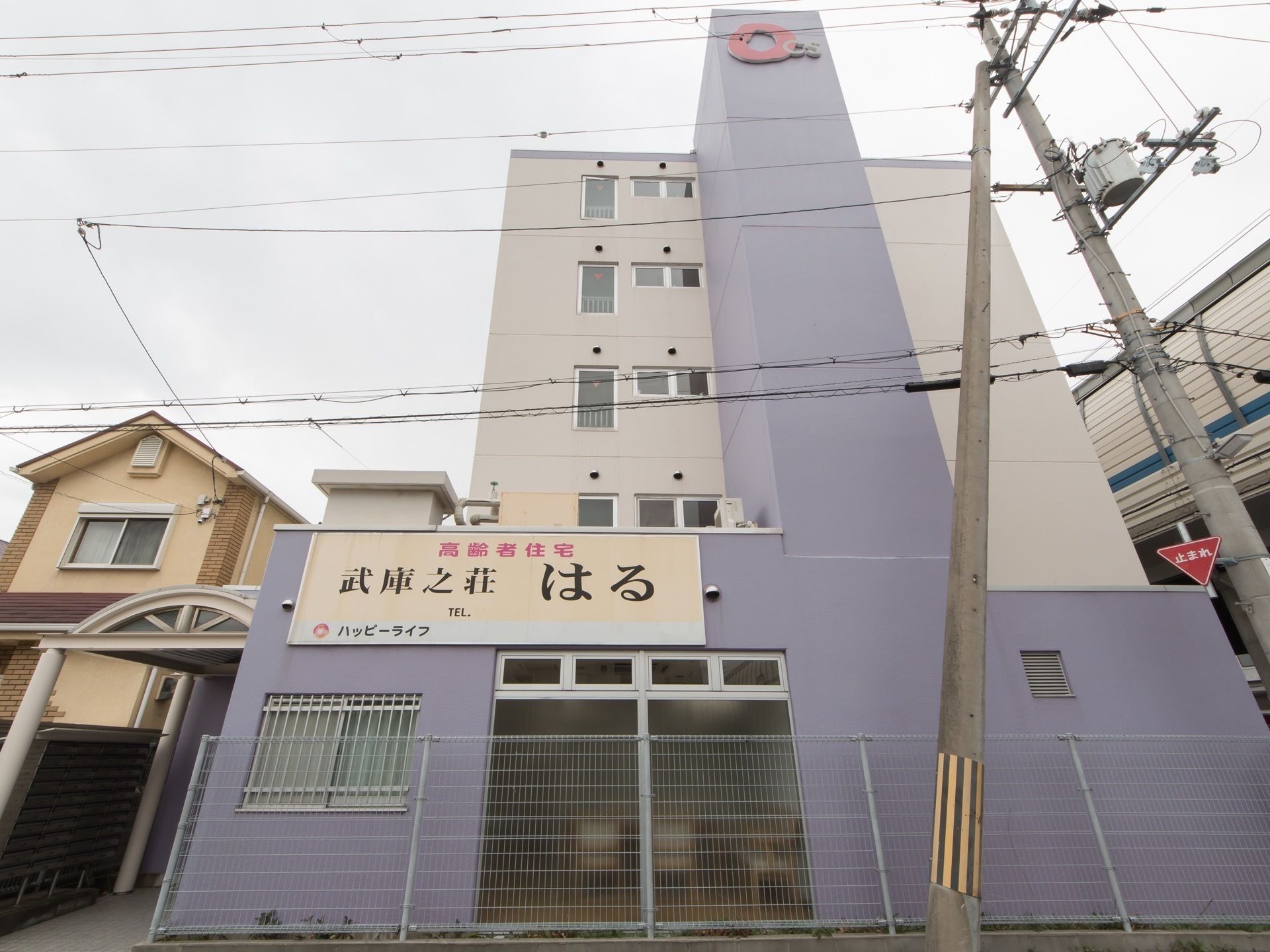 薄紫と白色のツートンカラーの外壁の建物で、手前の壁に大きな看板が設置されている。左側にアーチ型の屋根があり、その奥に玄関の入り口がある。