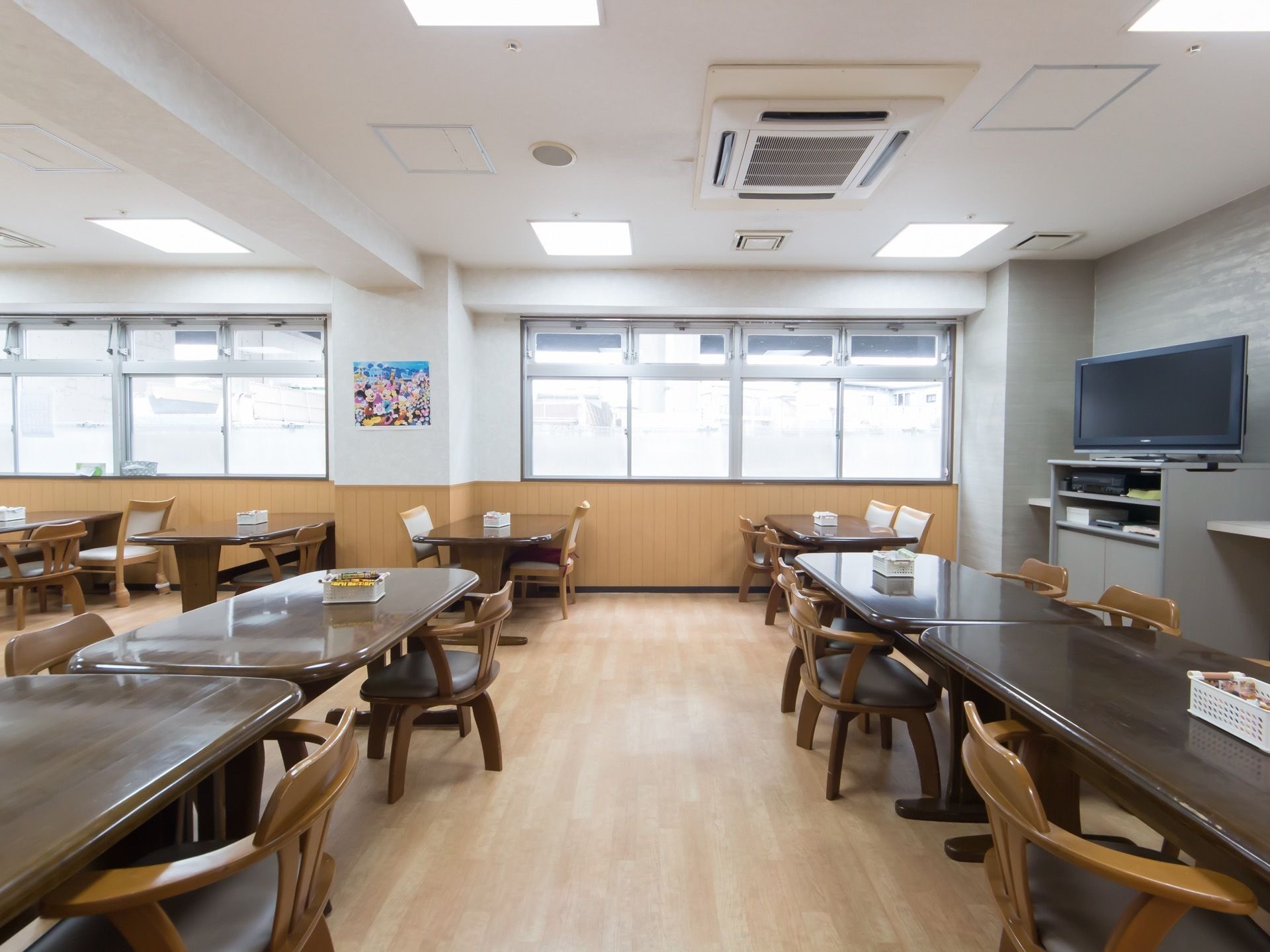 明るい色のフローリングの床の食堂で、ダークブラウンのテーブルがたくさん置かれている。右側の壁際にテレビが一台備わっている。