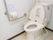 温水洗浄機能付きの洋式トイレである。壁にはトイレットペーパーホルダーとL字型の銀色の手すりが設置されている。