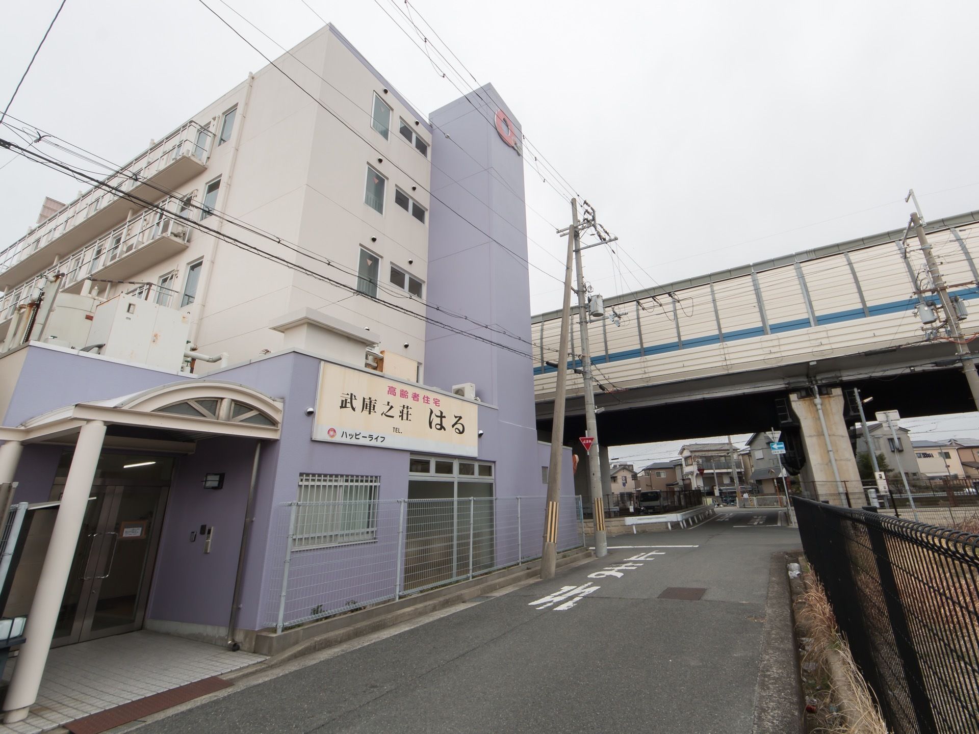 道路に面した外壁はすみれ色、奥は白色の5階建ての建物で、すぐそばには高速道路の高架がある。道路側の壁に、施設名の書かれた看板がある。