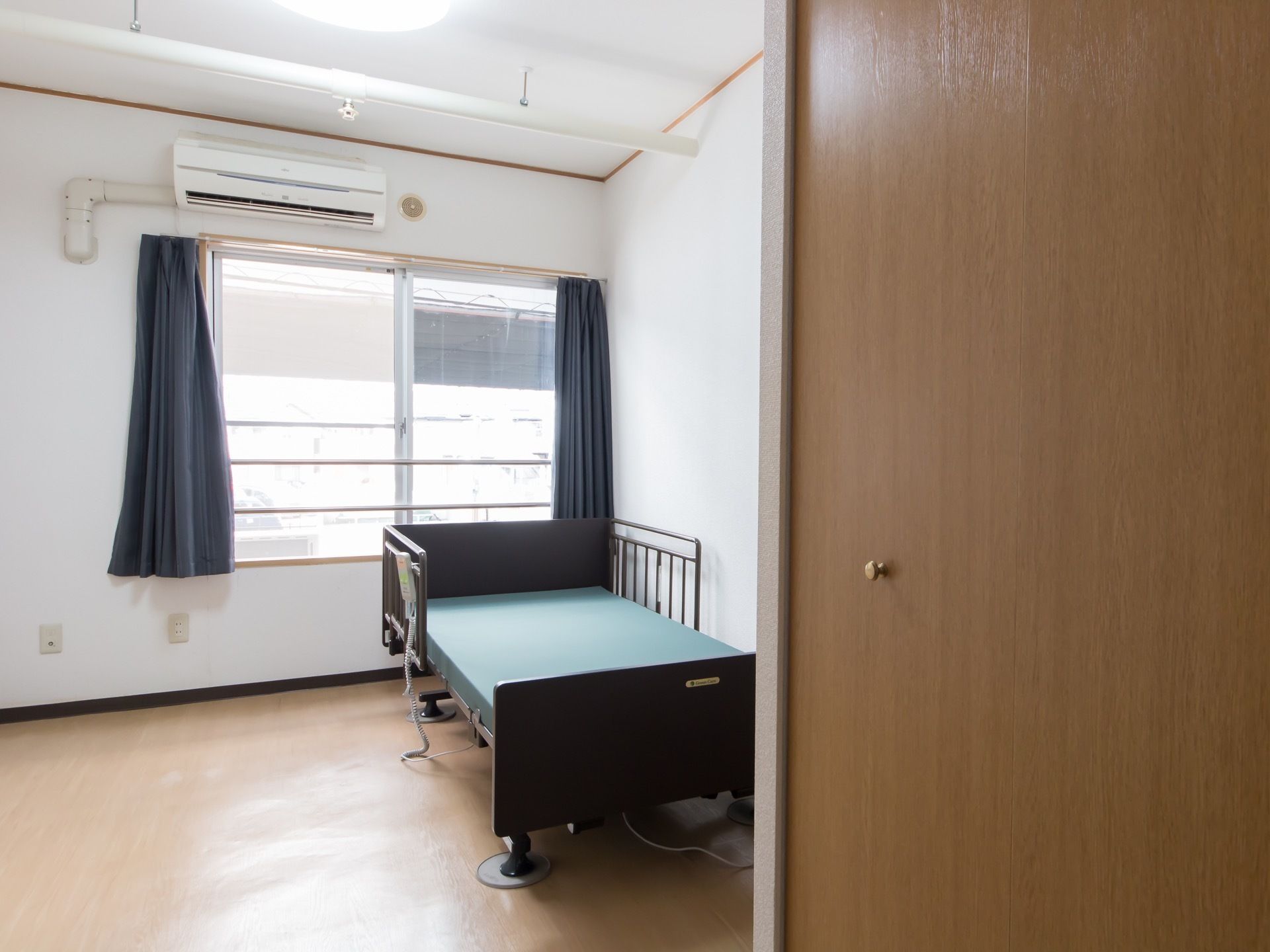 手前右側にクローゼットの扉があり、部屋の奥にベッドが一台置かれている。窓の上の壁に、エアコンが設置されている。