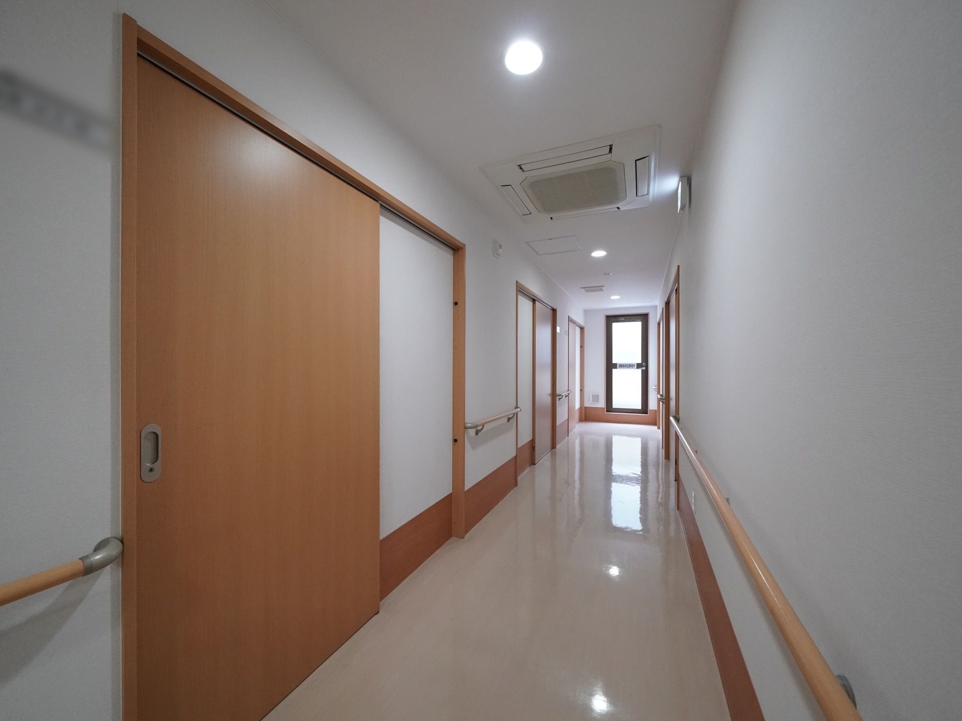 通路は白を基調とした空間で、突き当たりにある窓から日差しが差し込んでいる。両側の壁には木製の手すりが取りつけられている。