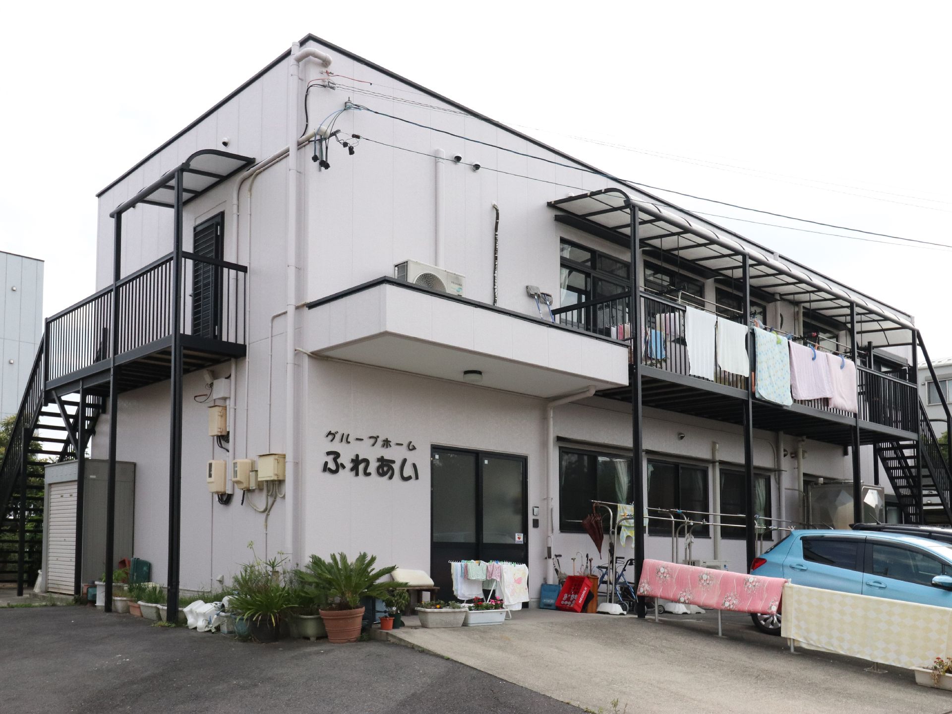 二階建ての建物の1階がグループホーム。引き戸の玄関。玄関前には沢山の鉢植えやプランターが置いてある。