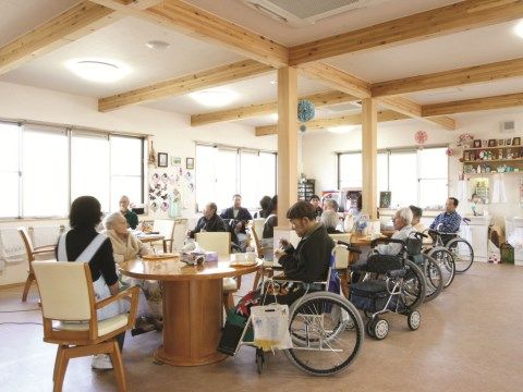 椅子や車椅子に座って過ごす人がいる、木製の柱や梁が見える白を基調とした食堂