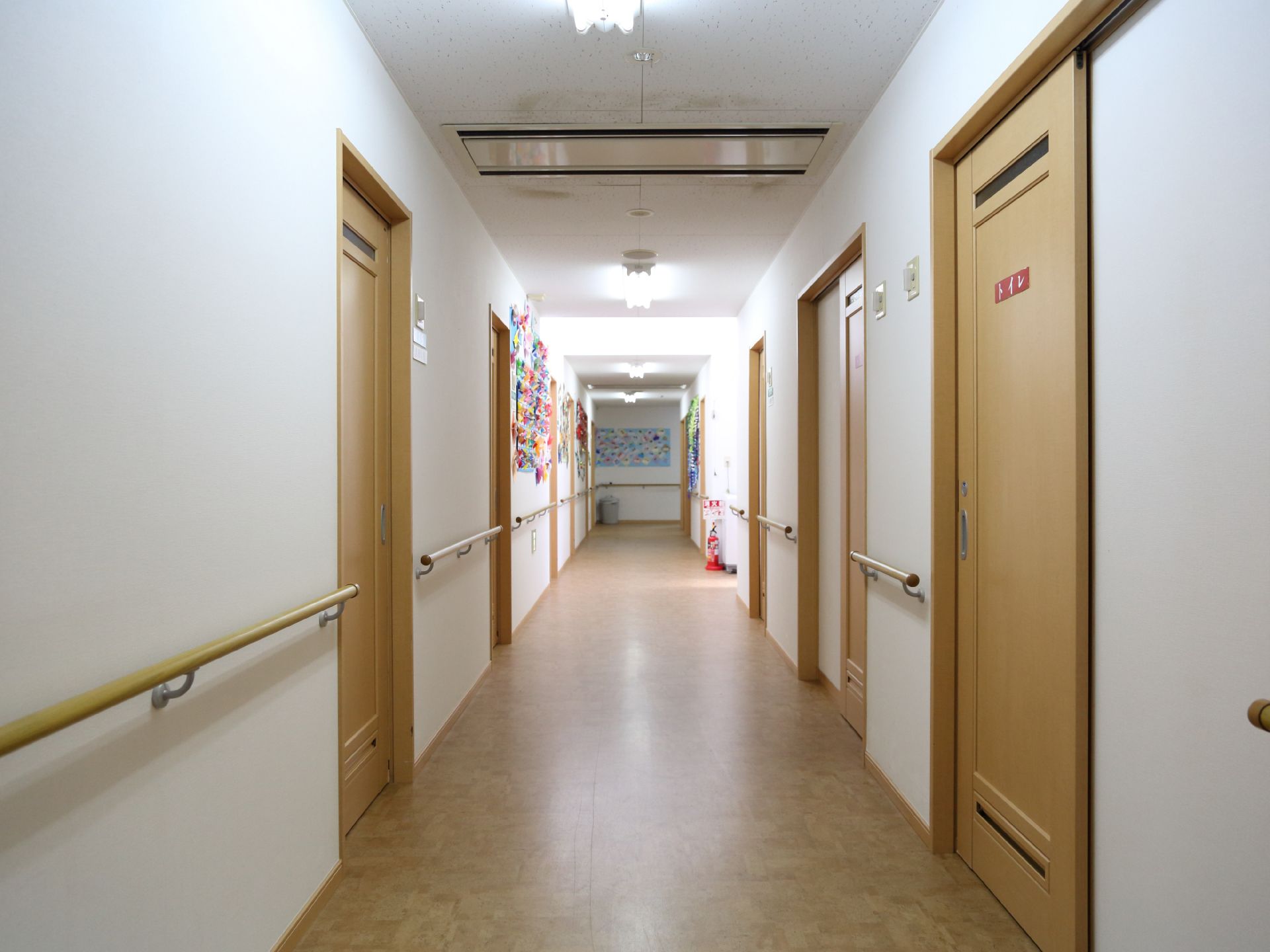 見通しの良い明るい廊下である。左右にはトイレや居室のドアが並んでおり、歩行補助の手すりが壁に設置されている。