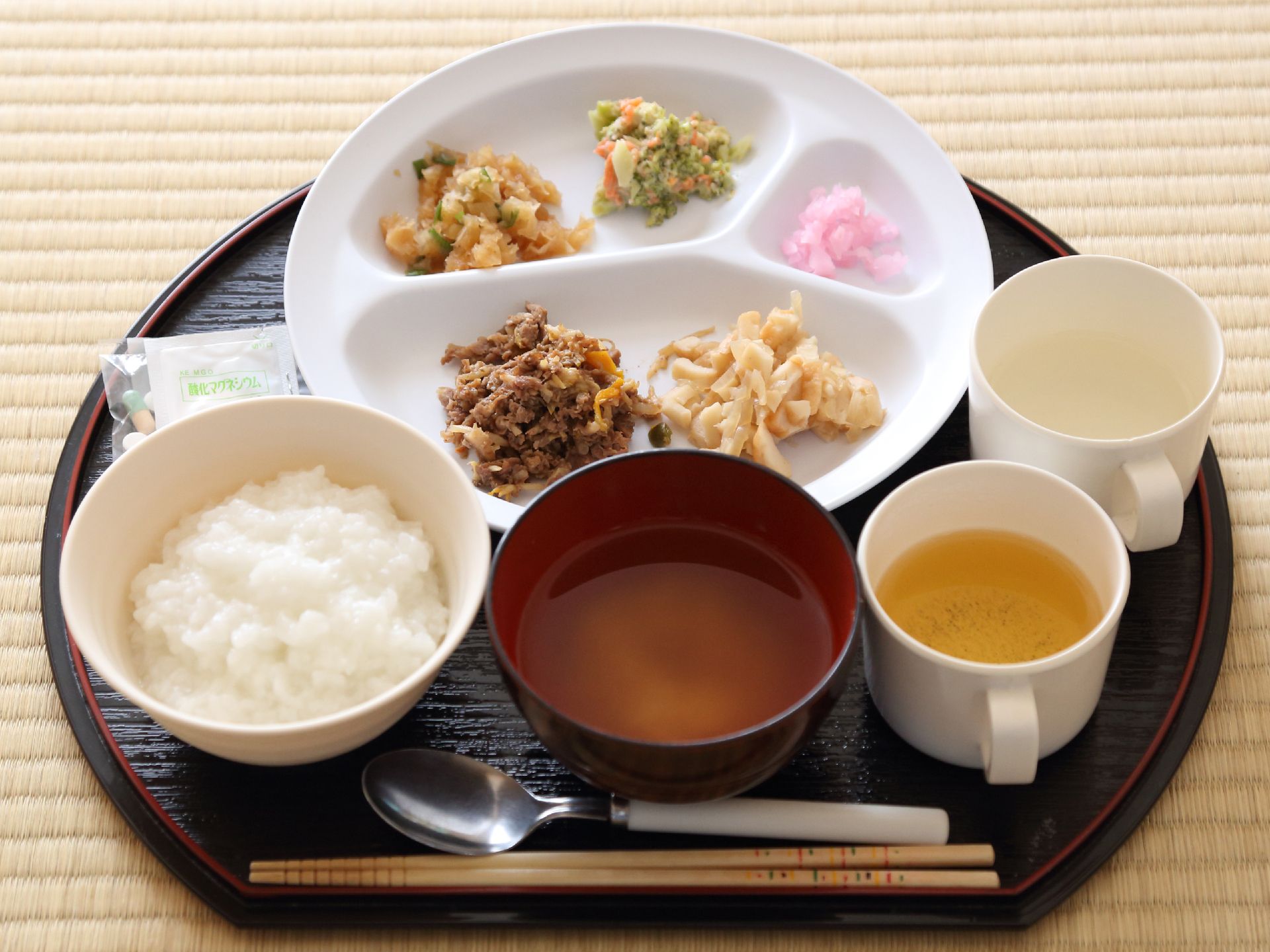 お粥やみそ汁、刻まれたおかずが黒い半月盆に置かれている。薬やお茶と水が入ったマグカップが用意されている。