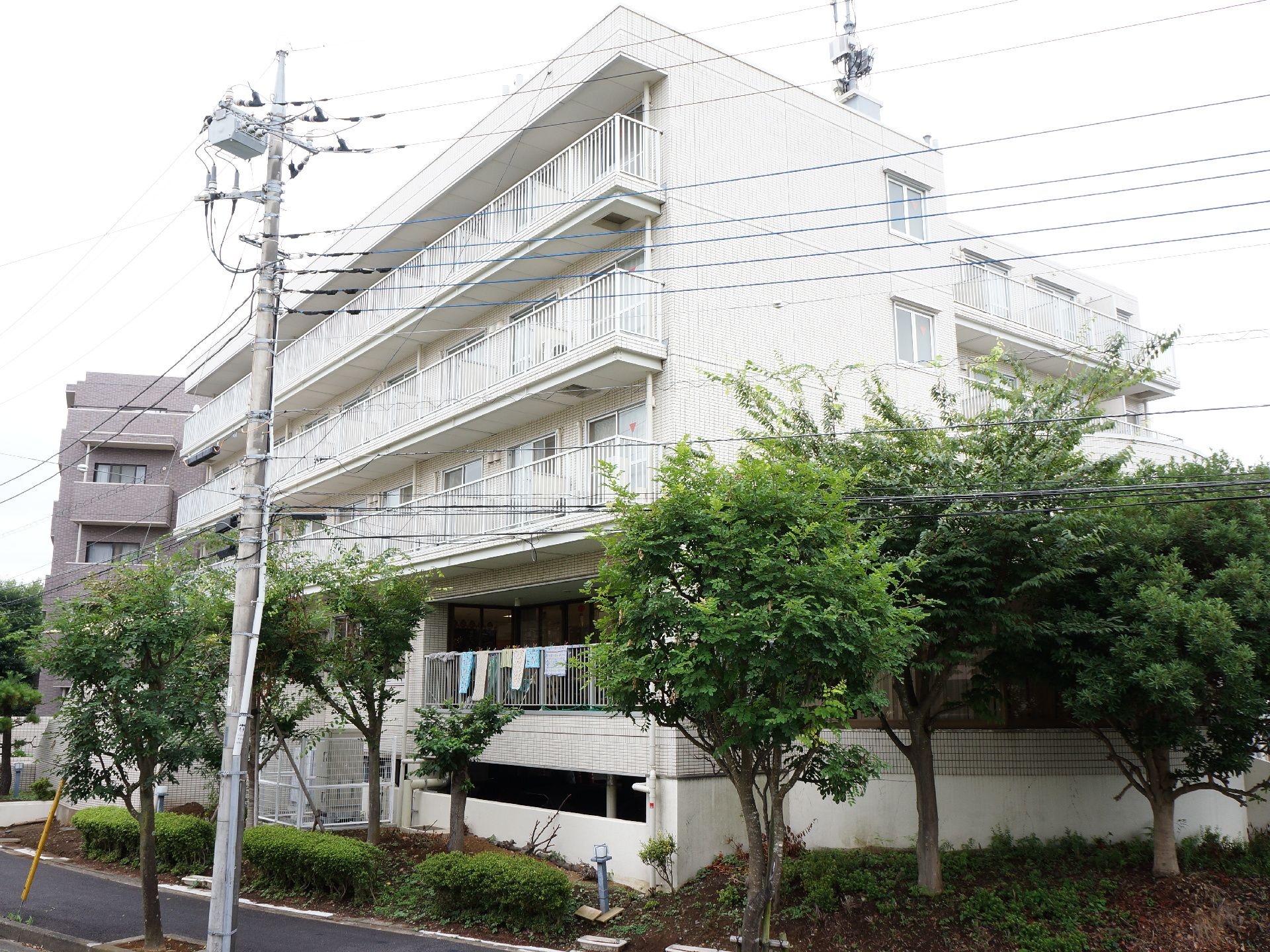 建物は白い5階建になっている。施設の周りには木が植えられている。施設前の道路は歩道の幅も十分に設けられている。