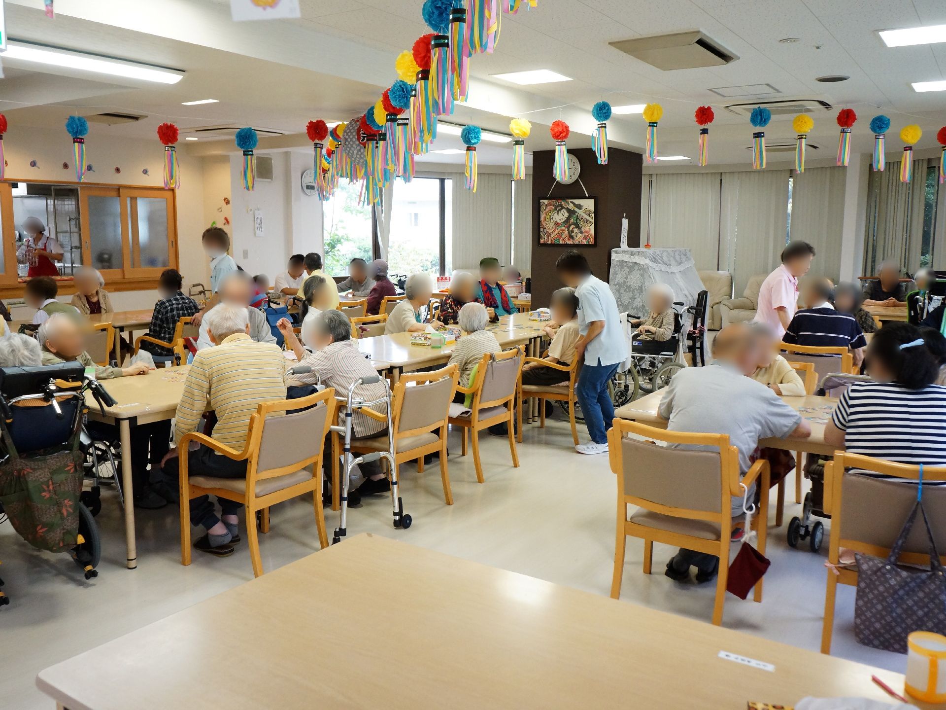 食堂は食事以外にも使用する。入居者様がレクレーションを楽しんでいる。沢山の机と椅子が配置されている。