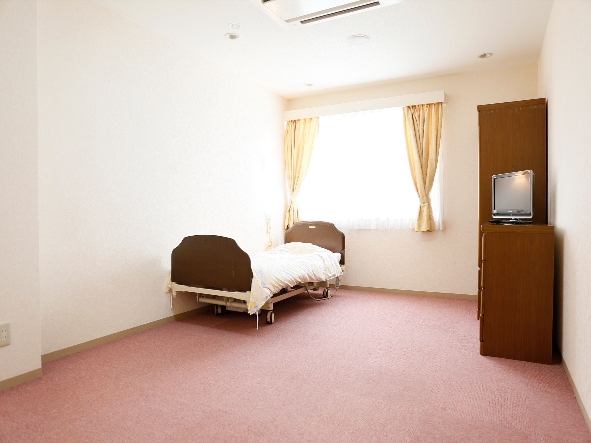 居室にはピンクのカーペットが敷かれており、ベッドやテレビ、タンスが設置されている。窓からは光が差し込んでいる。