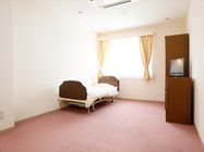 居室にはピンクのカーペットが敷かれており、ベッドやテレビ、タンスが設置されている。窓からは光が差し込んでいる。