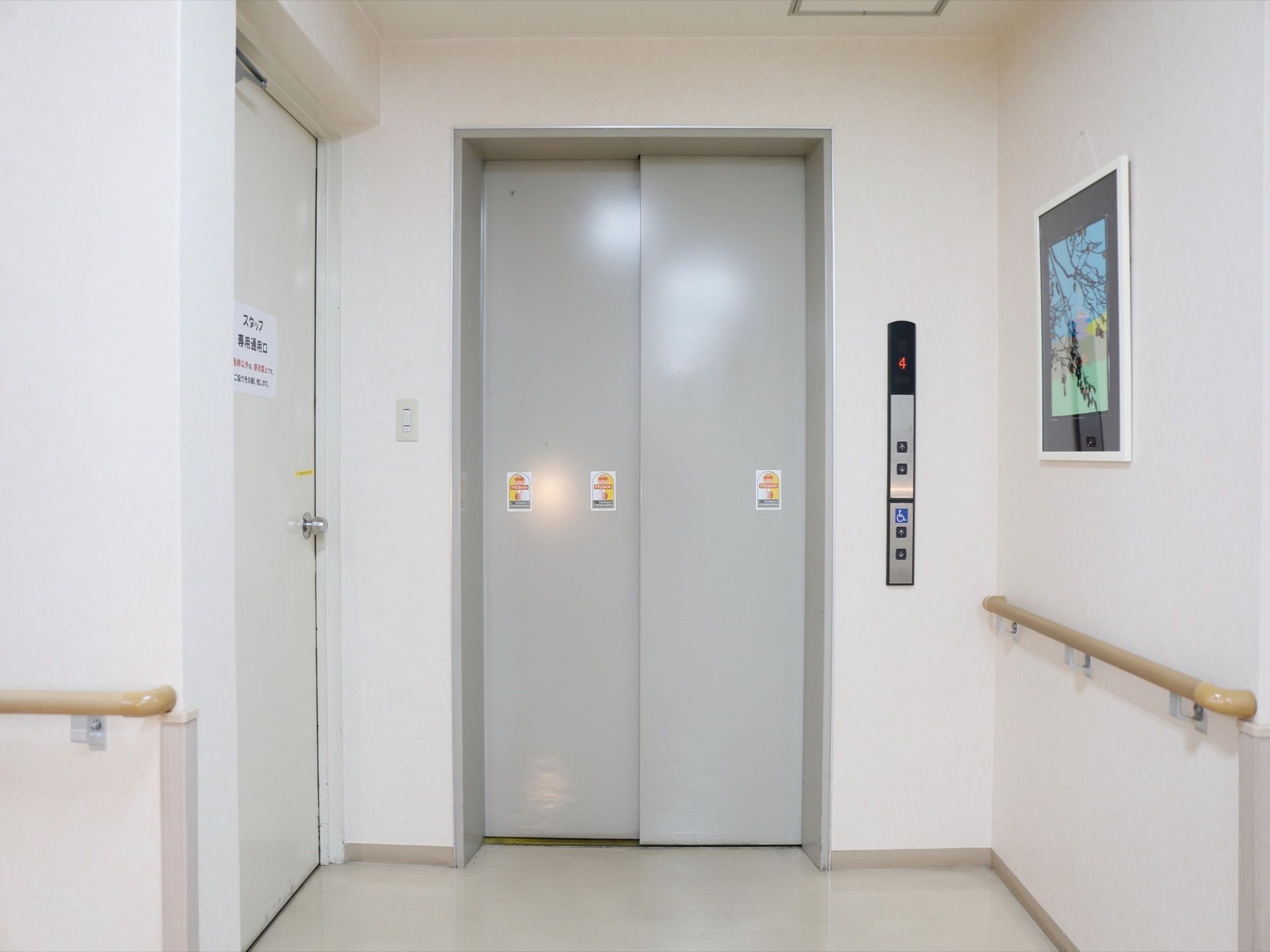 エレベーターは扉が閉まった状態となっており、表示板は4階で止まっている状態を示している。右の壁際には絵画が飾られている。