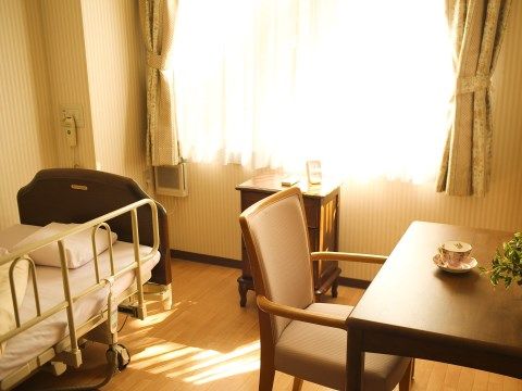居室には介護用ベッドが備え付けられている。窓際に小さな棚が置かれている。テーブルに椅子が１脚用意されている。
