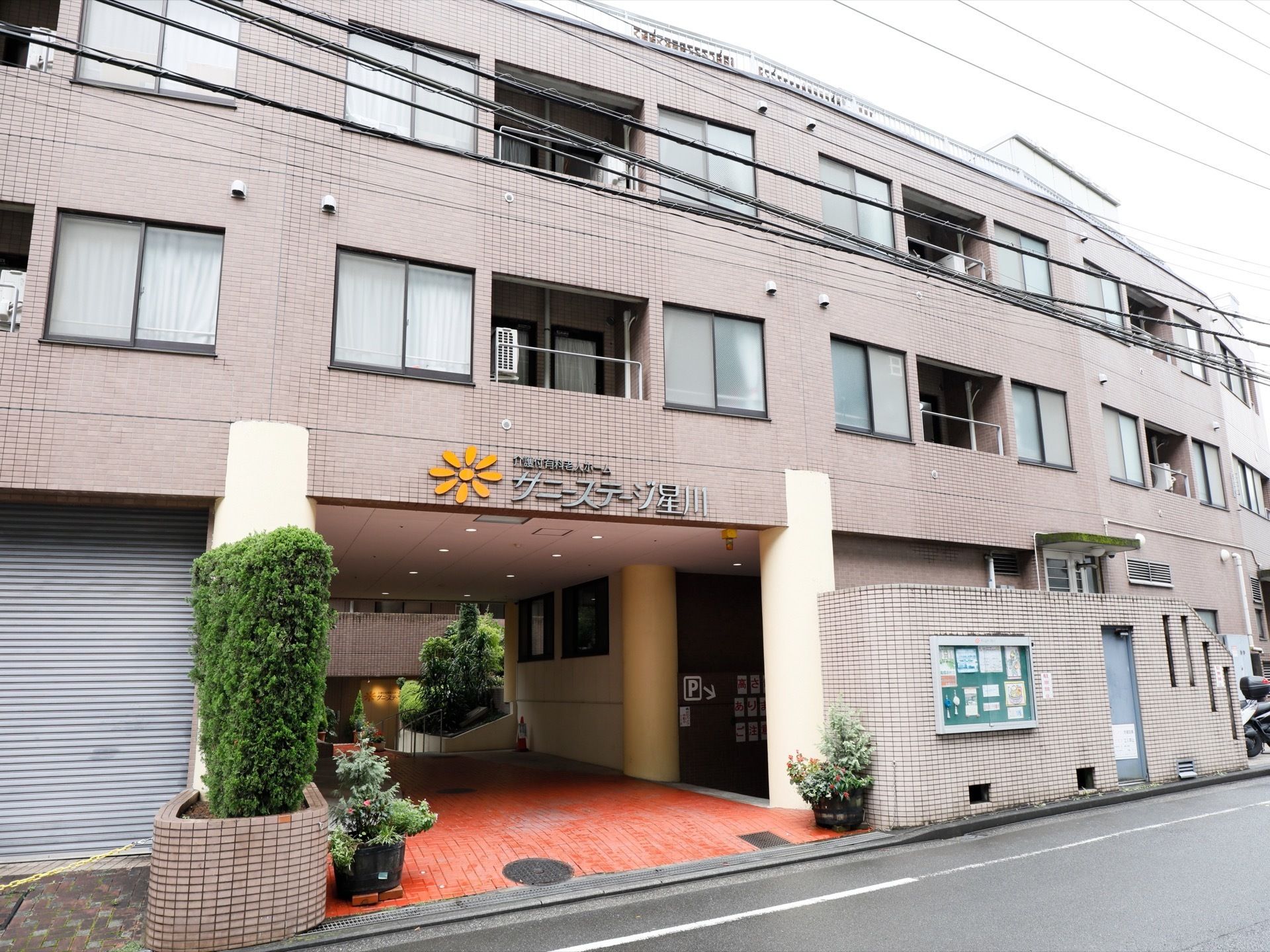 施設入り口は道路に面しており、両側には植物の植木鉢が置かれている。建物の壁はうす紫色で、掲示板の中にはお知らせなどが掲示されている。