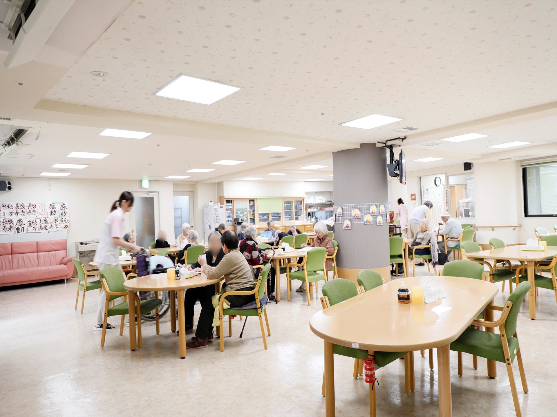 食事の風景。スタッフが入居者様に食事を配膳している。壁沿いにはソファが置かれ、近くに入居者様の習字の作品が飾られている。