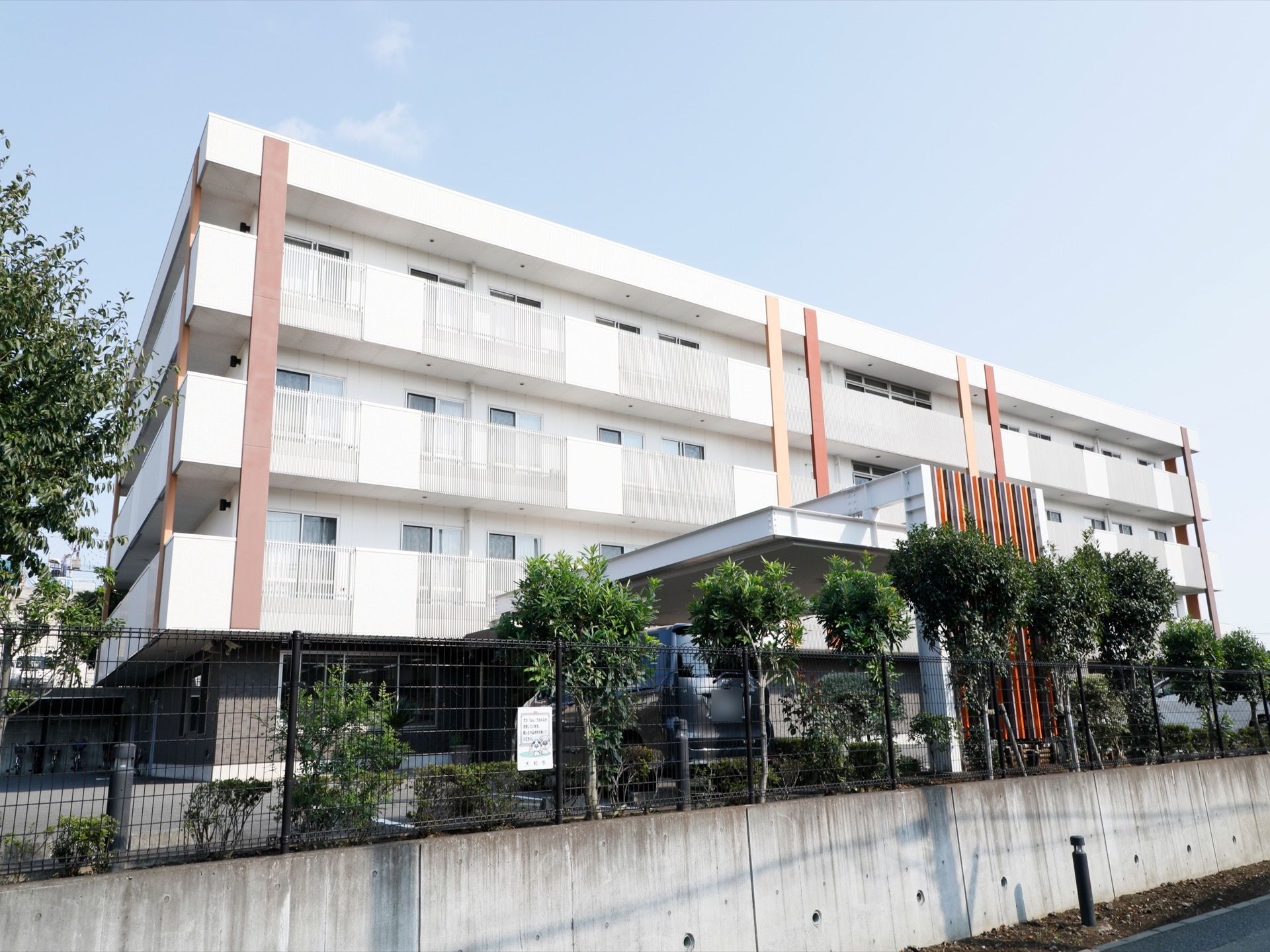 ４階建て施設は白い外壁で、広い敷地にある。敷地内に駐車場もある。ベランダがあり、マンションのような外観。