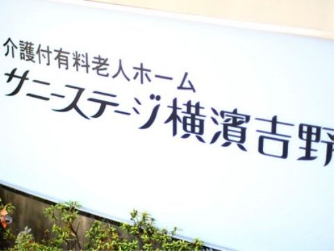 玄関には、白い看板で本施設名を標記している。施設は駅の近くにあるので、気軽にショッピングや小旅行に出かけることができる。