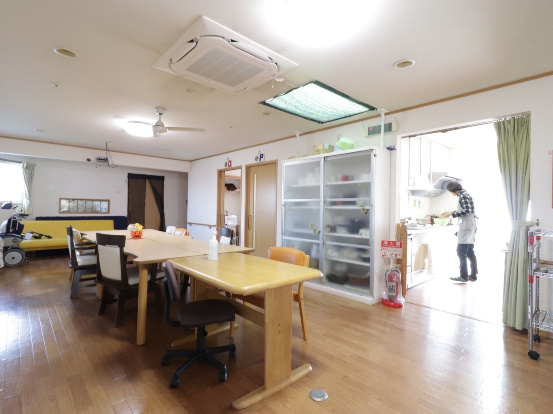 テーブルと椅子が並べられている。食器棚が置かれている。奥にはキッチンがある。壁際にはソファが置かれている。消火器が設置してある。