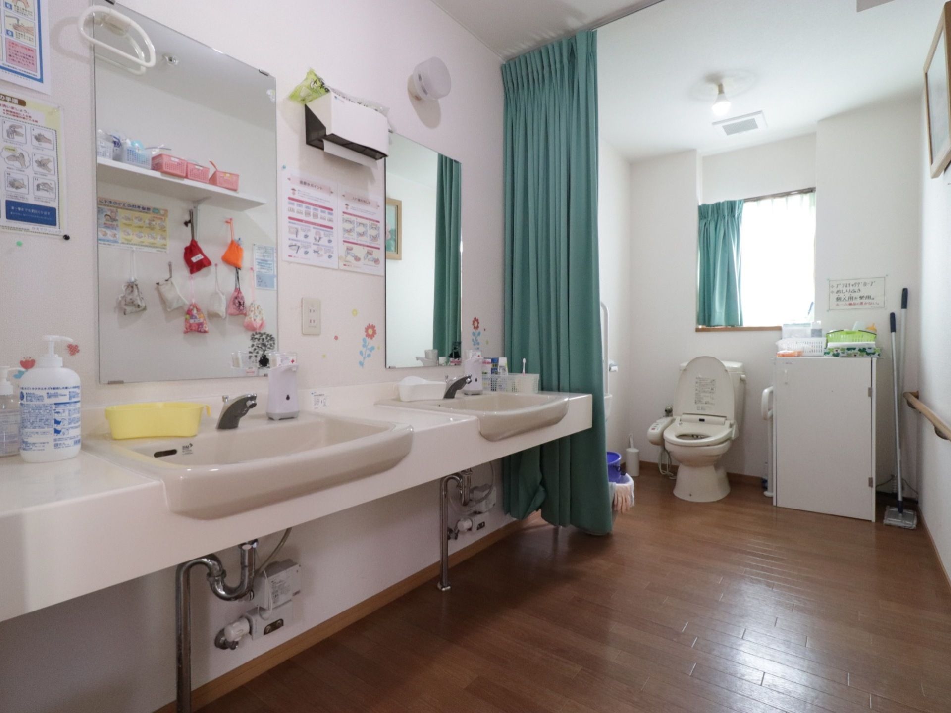 洗面台が２台あり、その奥にカーテンで仕切られたトイレがある。洗面台はバリアフリータイプである。センサー式のハンドソープが置かれている。