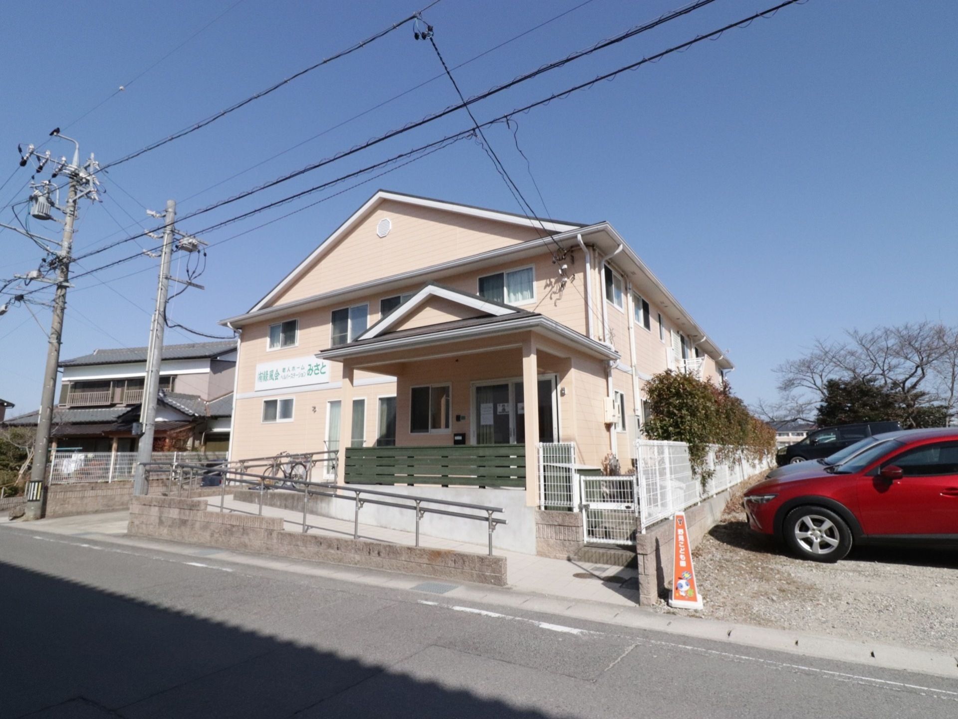 施設は淡いベージュ色の外壁の木造２階建てである。玄関までスロープが設置されている。スロープには手すりが付いている。