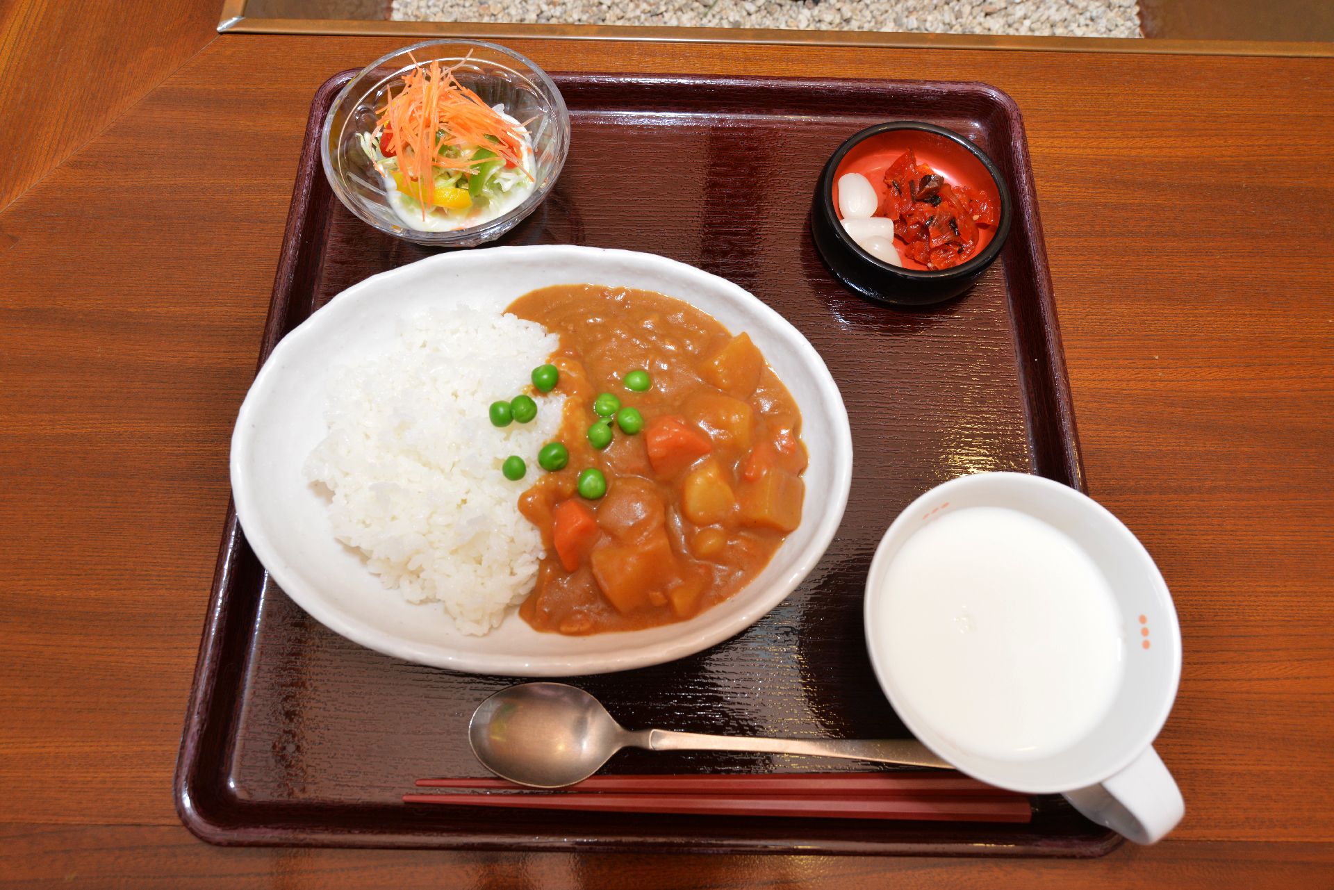 カレーライスとサラダ、福神漬などが茶色のトレーに並んでいる。牛乳も添えてある。野菜がたくさん入った家庭的な雰囲気のカレーである。