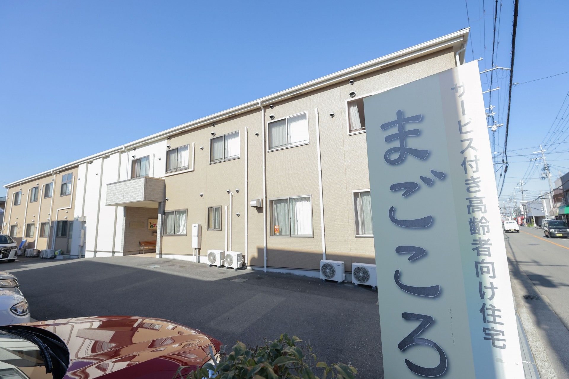 施設の出入口には、施設名の書かれた看板が立っている。看板の周辺には樹木が植えられたスペースがあり、その横には駐車場がある。