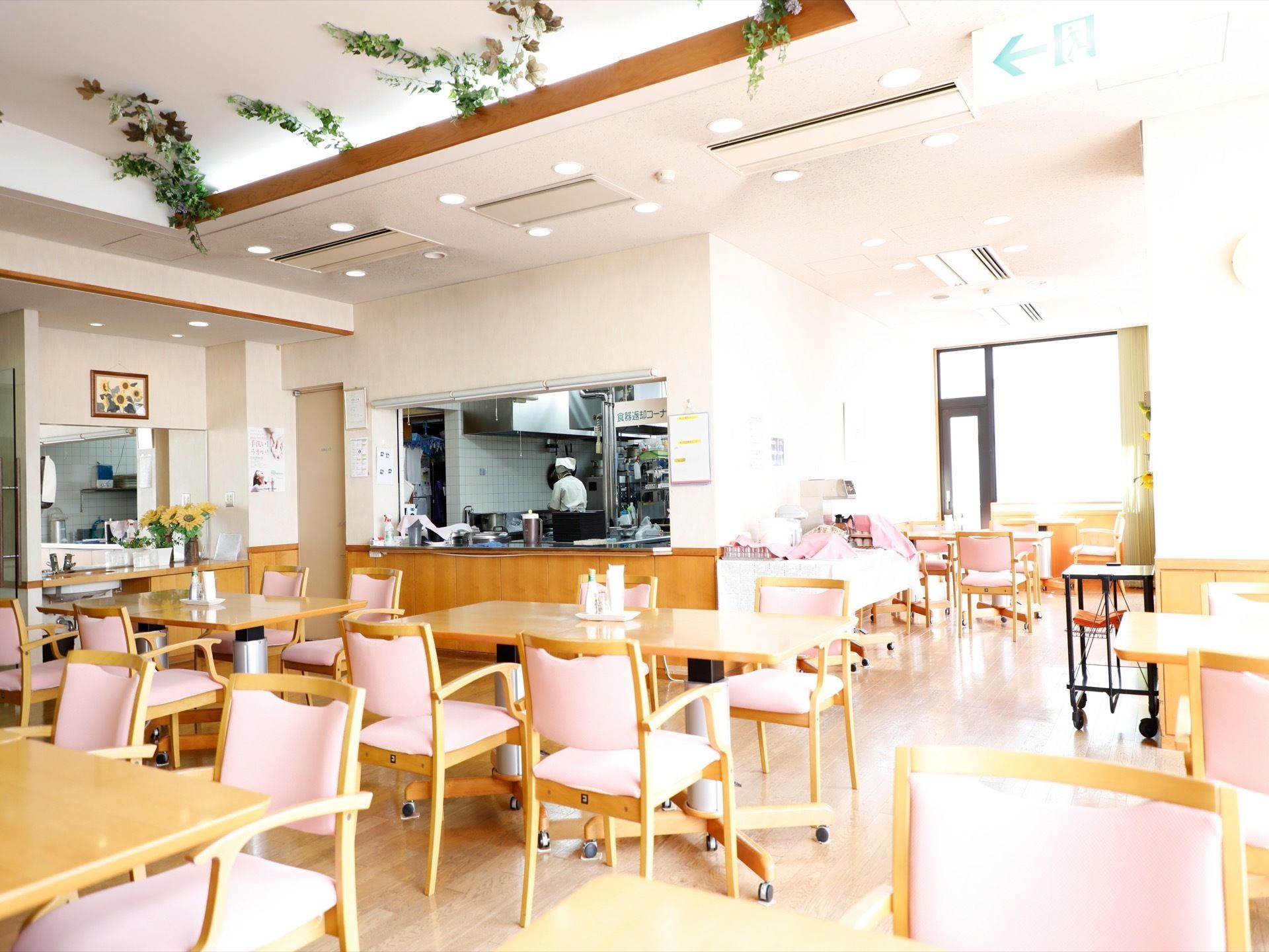 沢山のテーブルと椅子が置かれている。左奥には配膳の窓口があり、その左側には洗面台がある。天井には草の飾りが付いている。