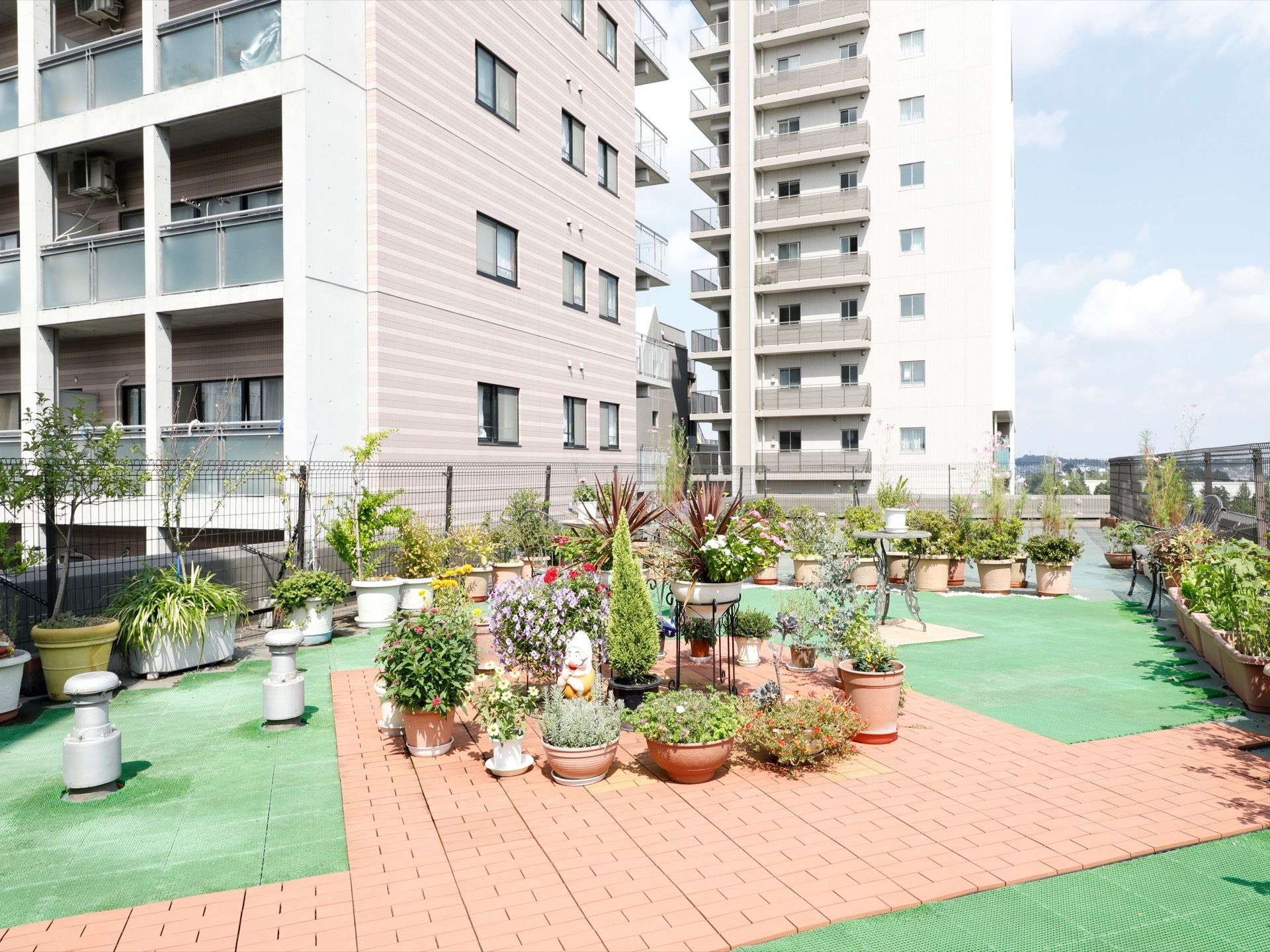 床にはレンガや人工芝が敷かれている。中央やその周辺には鉢に入った植物などが沢山置かれている。右側や左側などには柵がある。