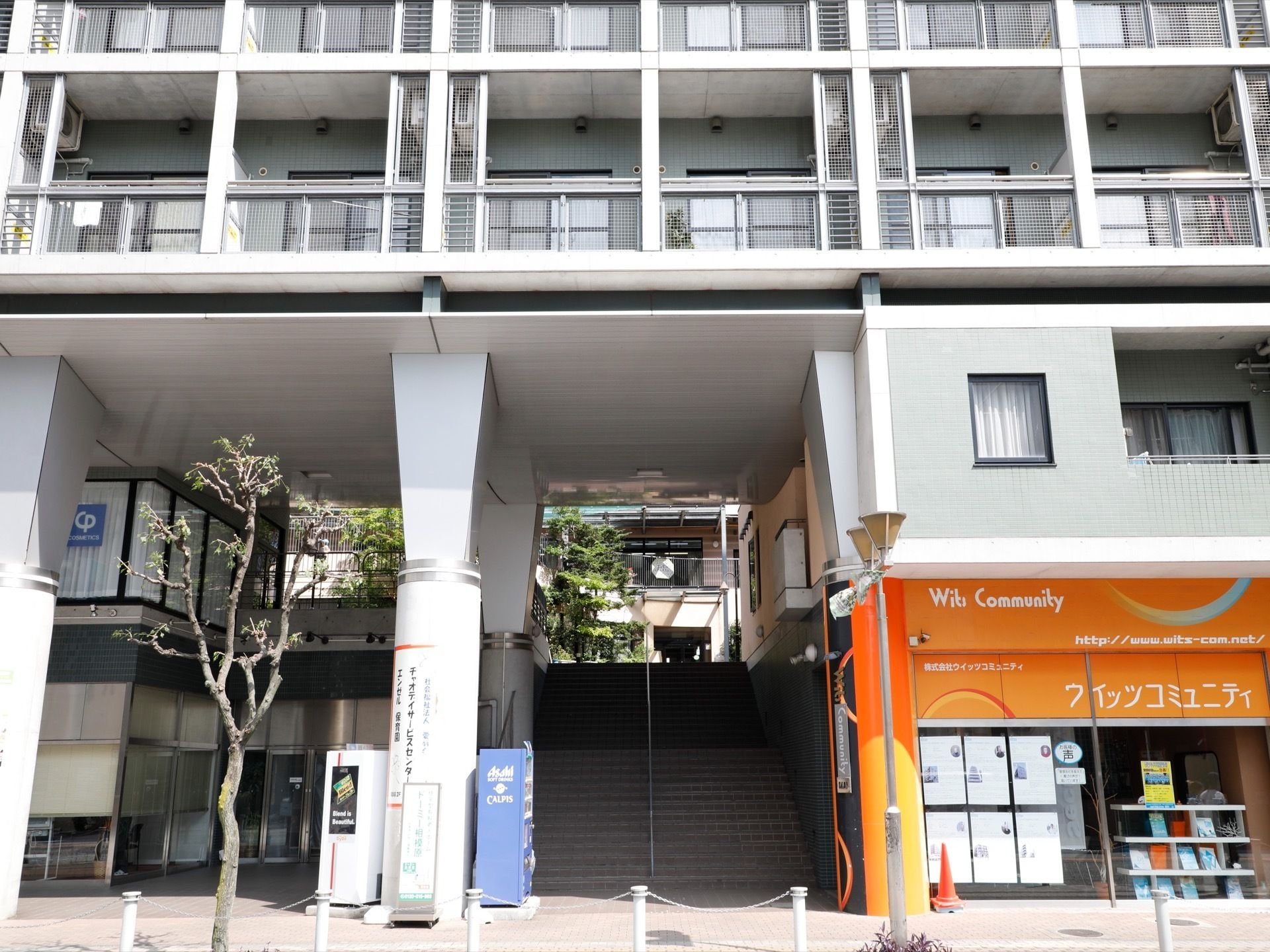 建物の前は道路と遊歩道がある。下の階にはテナントが入り、上が住居になっている。１階部分には自動販売機もある。