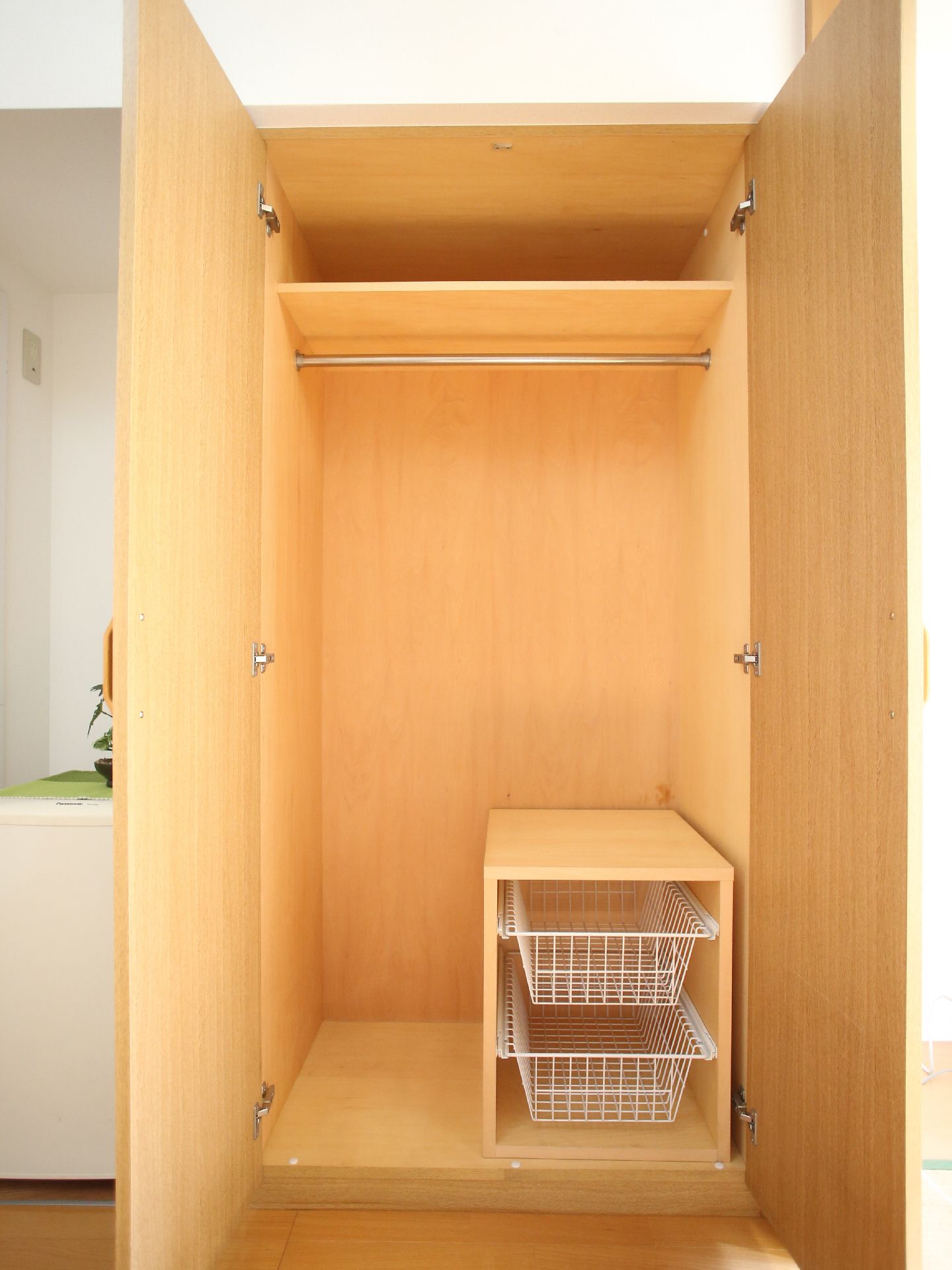 木目調の両開きの収納庫である。内部も木目で上部に棚があり、すぐ下にポールを備えている。下の横側にボックスを作り、中に２つの籠を入れている。