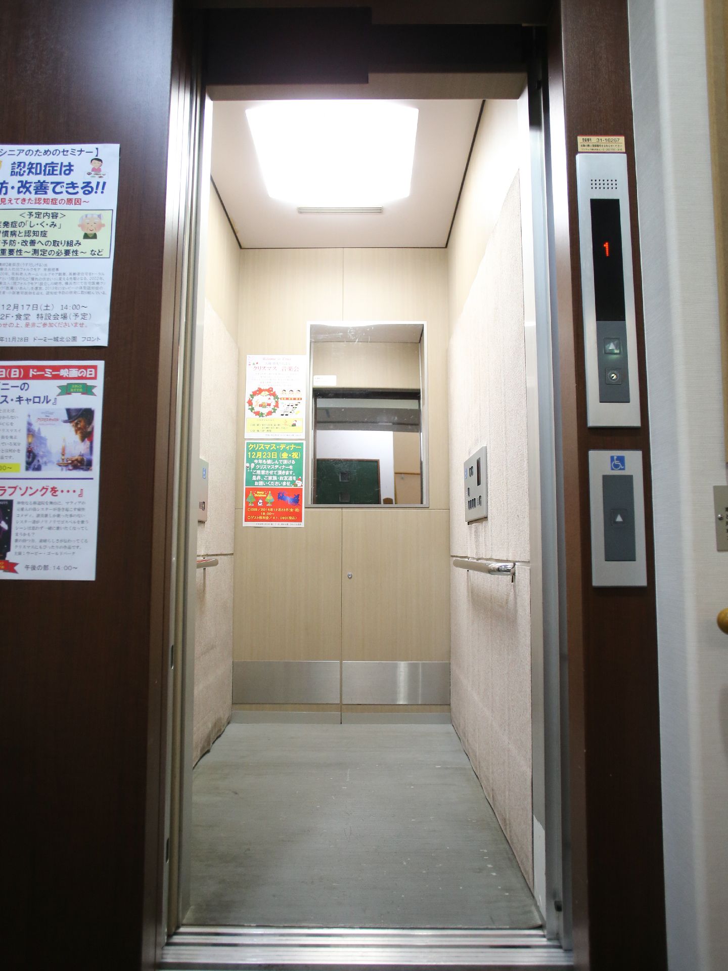 奥行に長いエレベーター内である。正面には鏡があり、両側に手すりを備えている。鏡の横には、広告などをはっている。