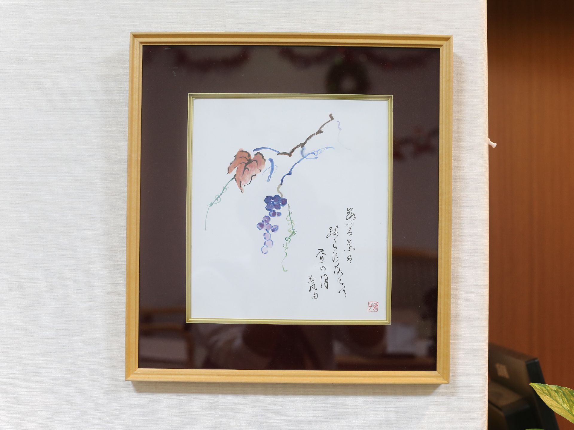 焦げ茶色に黄土色の枠のある額の中央に、ぶどうの実と葉が描かれ、横下に詩が添えられた絵を白色の壁際に掛けている。