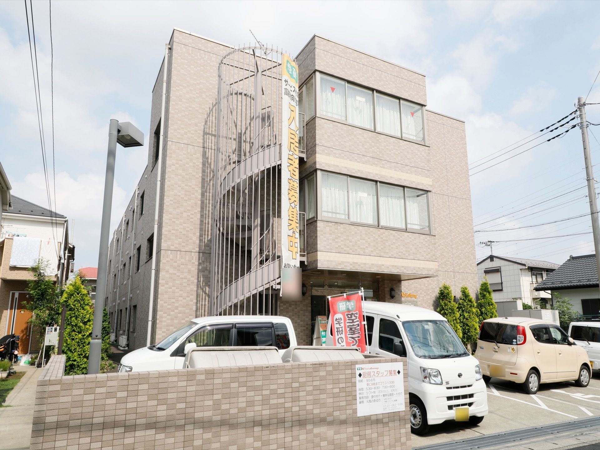 ブラウンを基調とした3階建ての落ち着いた建物。正面には螺旋階段が設置されている。玄関の前には車が数台停まっている。