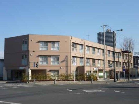 住宅街の中にある、明るい茶色の建物。手前に大きな道路がある。入り口の上には施設名が書かれており、周りに花壇がある。