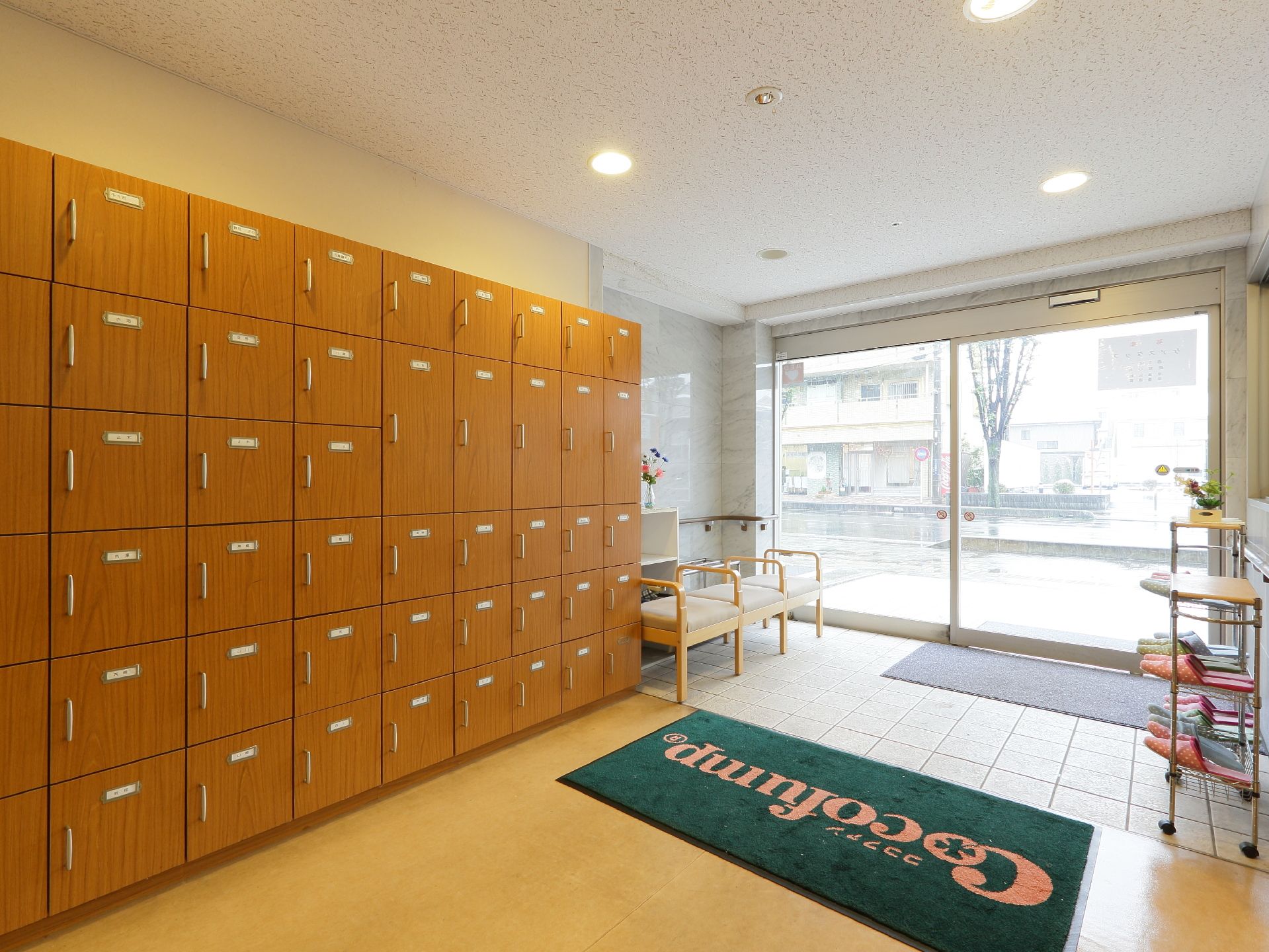 広く開放されるガラスの自動ドアから光が差し込む明るい雰囲気の玄関。扉付のシューズボックスが整然とならび、着脱用の椅子が置かれている。
