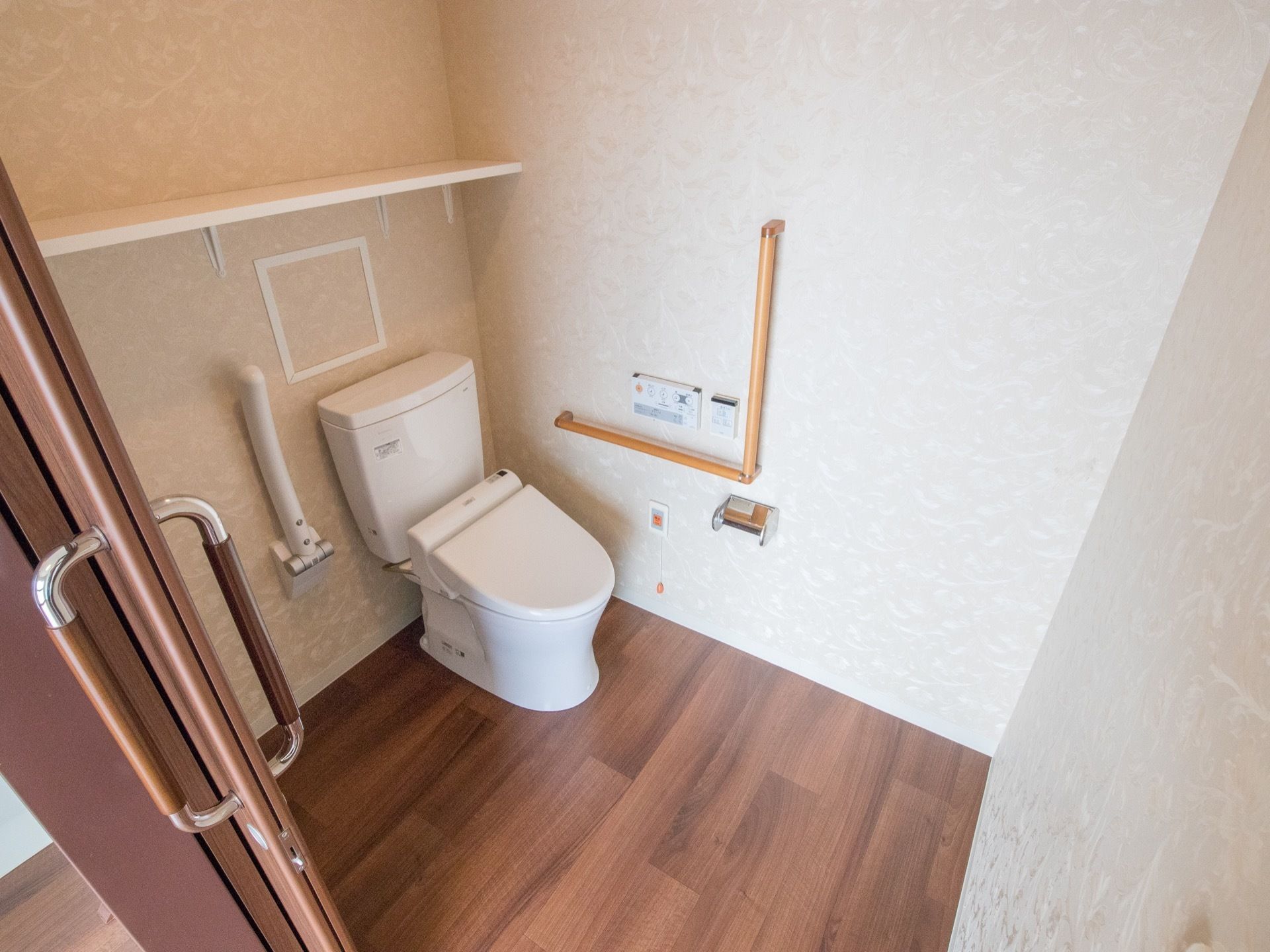 内装は細かい模様が入った淡い色の壁紙である。白いタンク付きの洋式便器の横に可動式の手すりが設置されている。