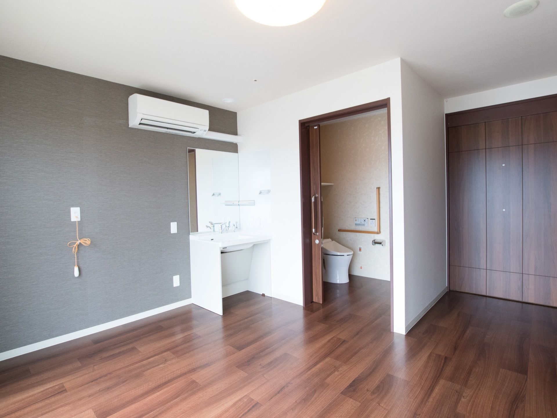 壁に設置されたエアコンの下に洗面台がある。トイレのスライドドアは全開になっていてい、中の白い便器が見える。