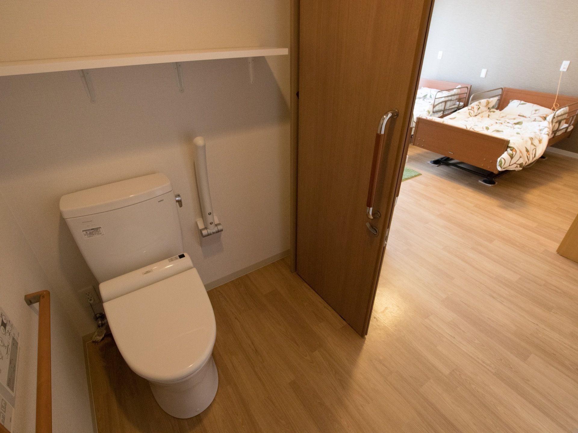 トイレの扉が開かれ、居室の転落防止柵が取り付けられたベッドが見える。洋式便器の上の壁に棚が設置されている。