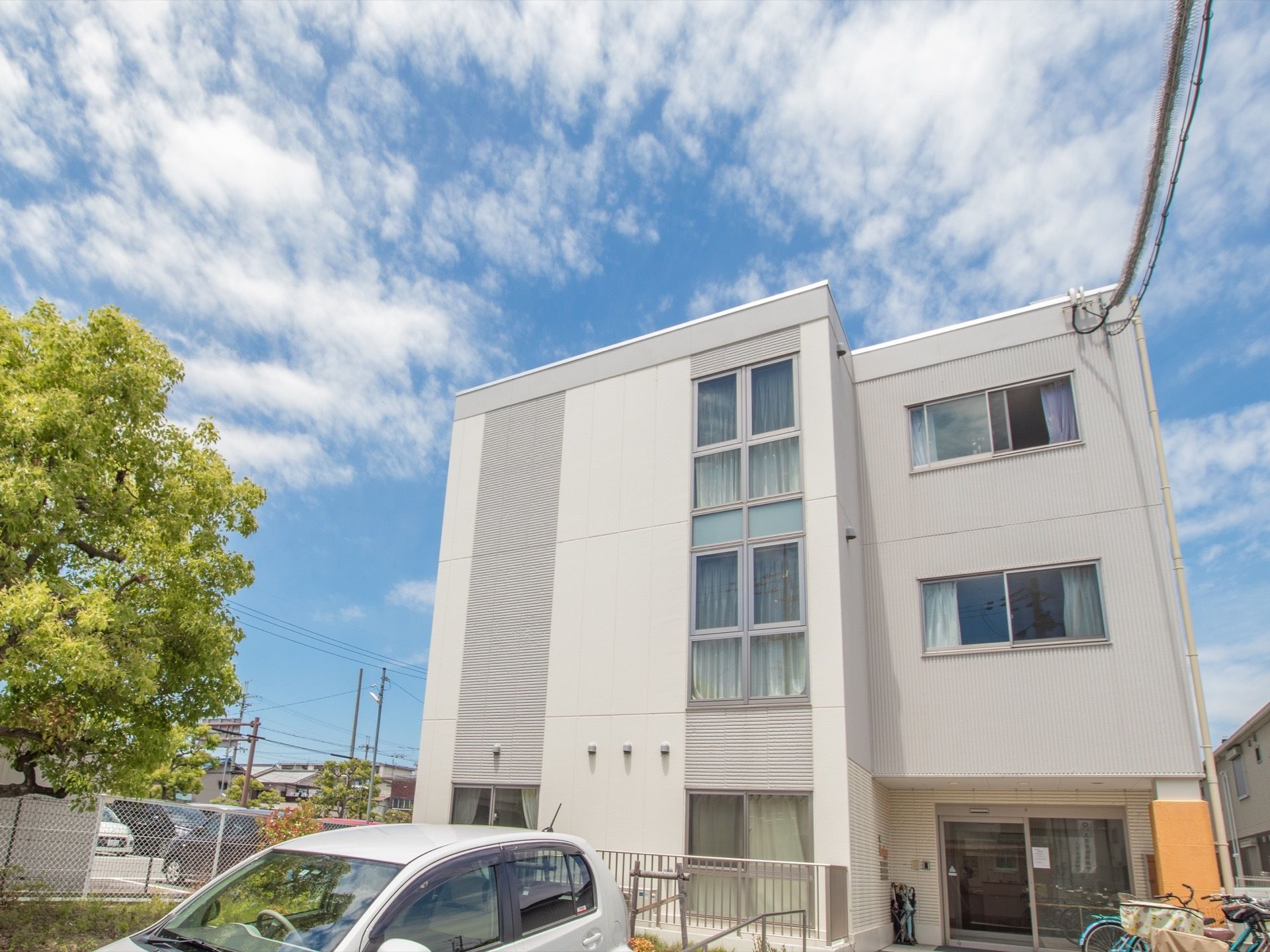 雲がかかった青空の下に、白とグレーの３階建ての建物がある。建物の背後に民家が建ち並び、車が停められている。
