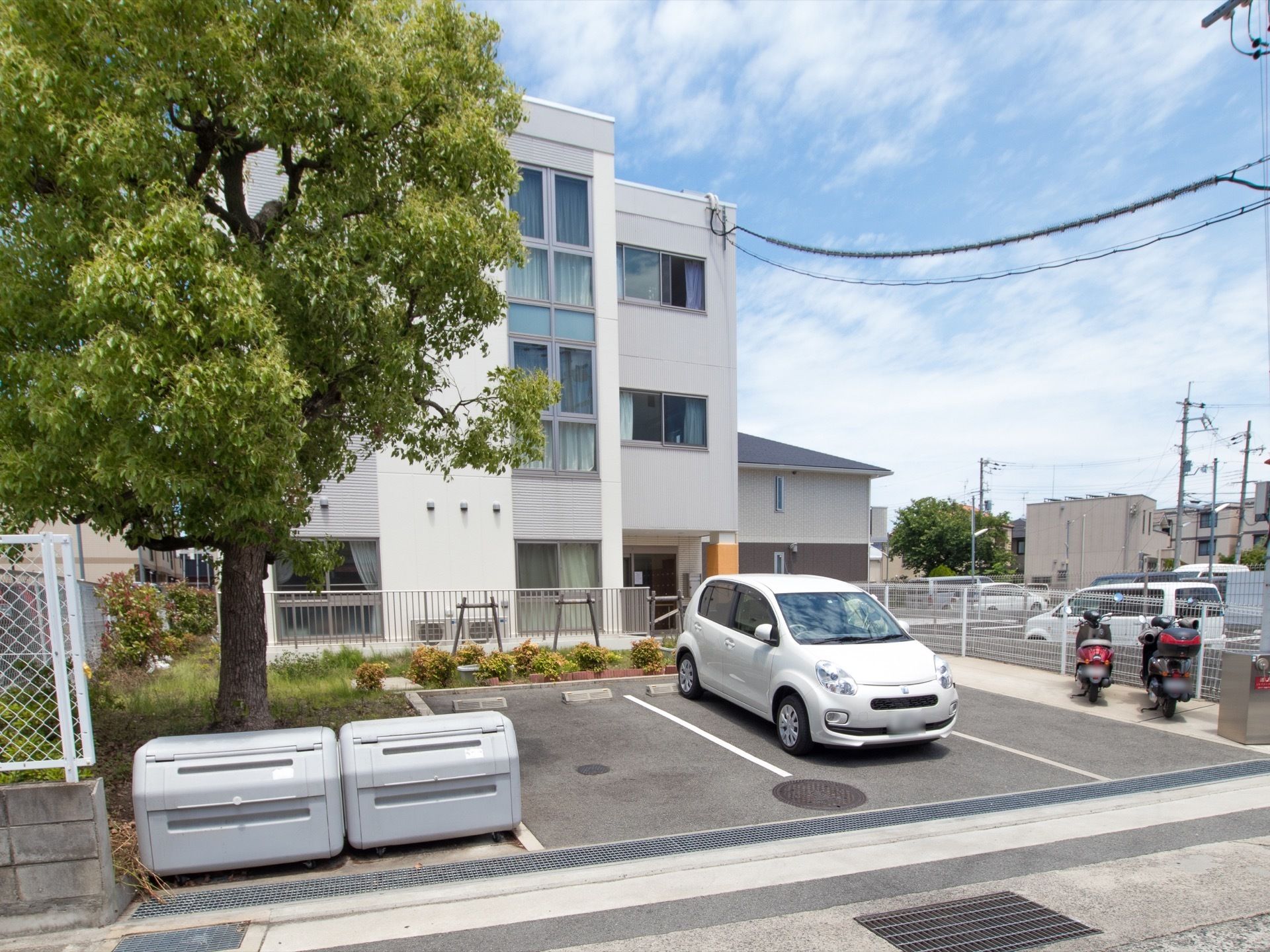 白い建物の前にアスファルトで舗装された駐車スペースがある。大きな木の下にダストボックスが２つ並べて置いてある。