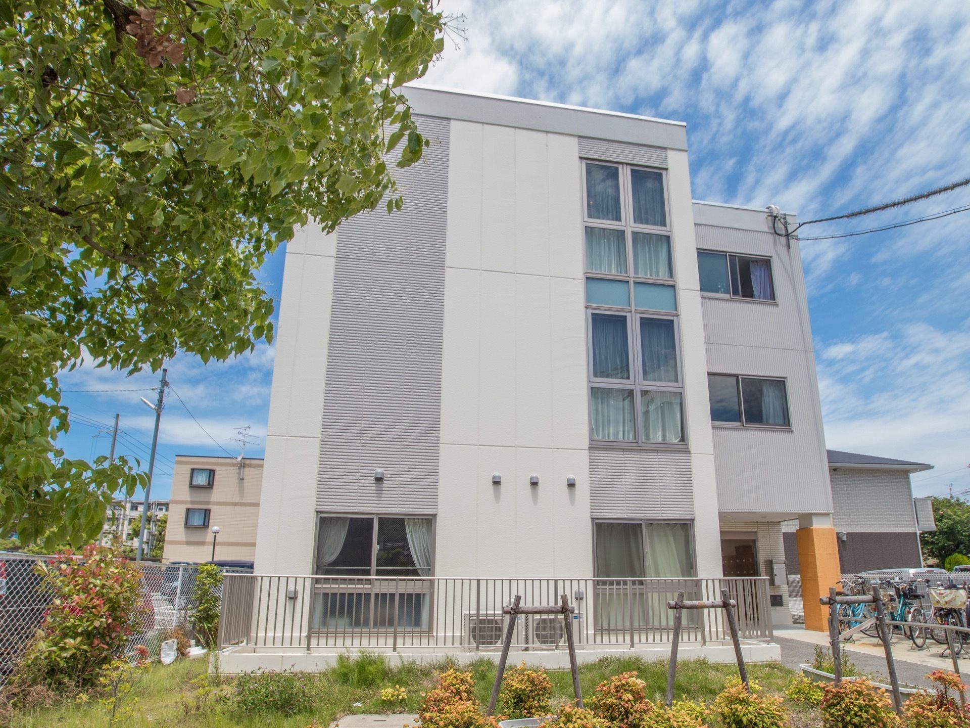 スカイブルーが美しい青空にすじ雲が掛かっている。３階建ての白い建物の前に大きな木がそびえ立っている。