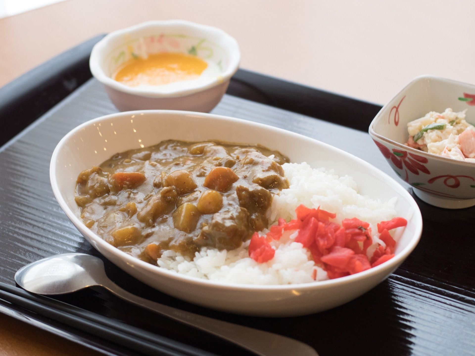 黒い配線盆にカレーと２種類の小鉢とスプーンが置いてある。カレー皿に福伸漬けがのった白米と具だくさんのルーが盛り付けられている。