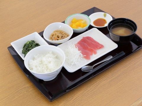 盆の上に、ご飯、みそ汁、マグロの刺身、葉物、レンコンの煮物、黄桃のデザートがそれぞれ皿に盛りつけられている。