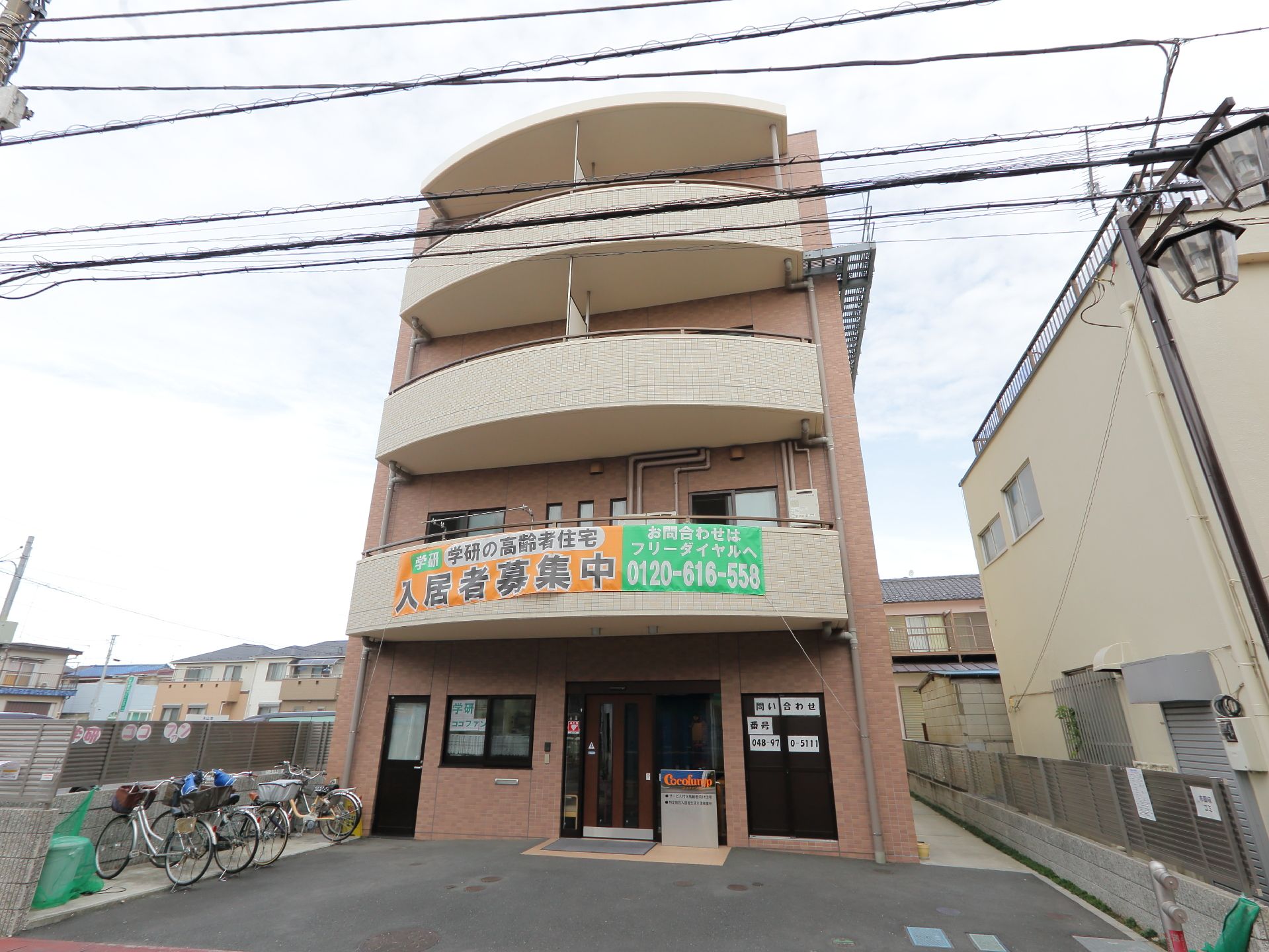 施設の玄関近くでは自転車置き場がある。大きく「入居者募集」のポスターが掲示されている。薄い茶色の建物で4階建て、2階以上にはベランダがある。